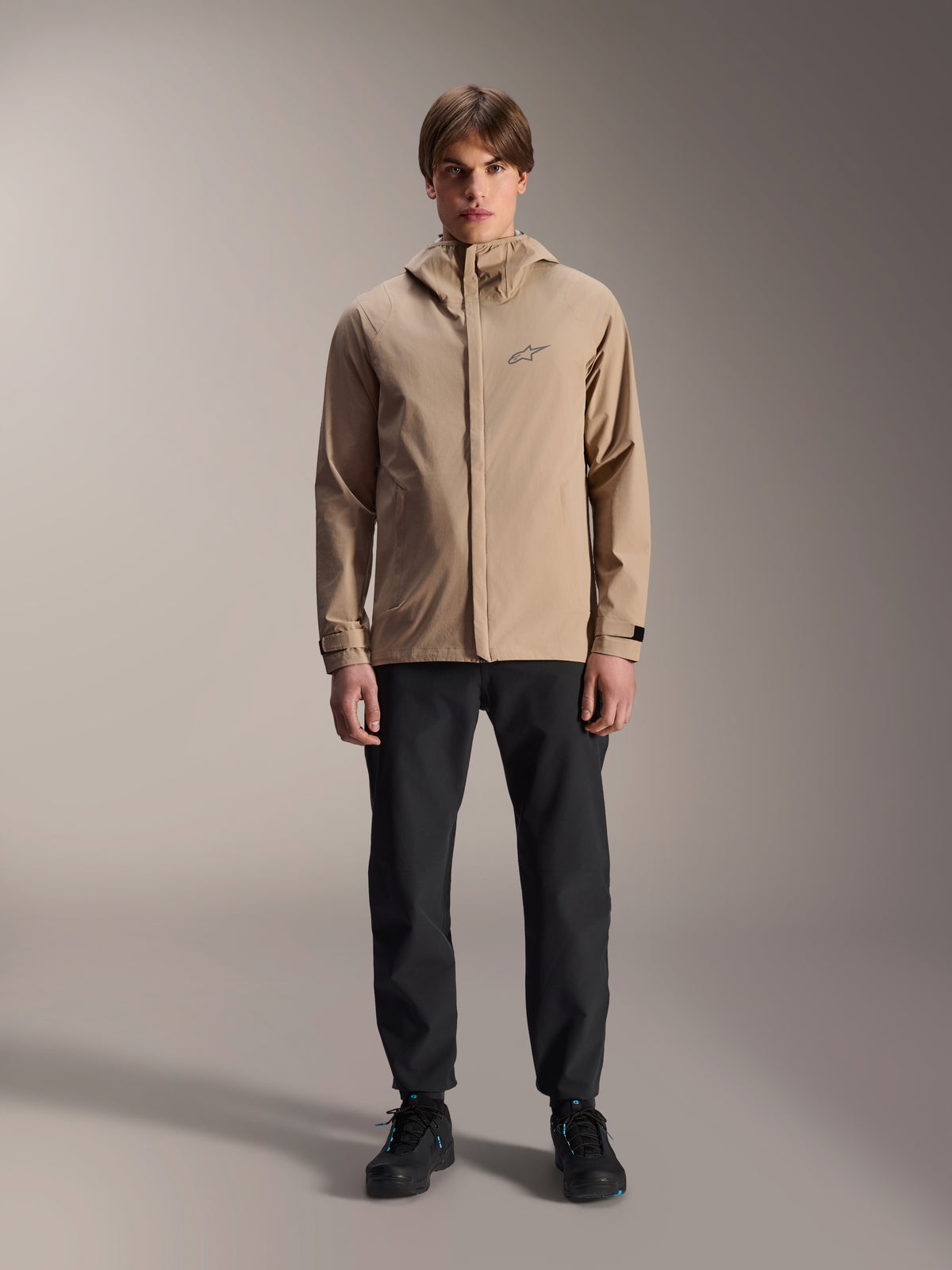 Alpinestars A-Dura Rain Jacket, lysebrun cykeljakke, båret med sorte bukser og sorte sko, med et lille brystlogo og hætte, professionelt mountainbike-tøj til kørsel i al slags vejr.