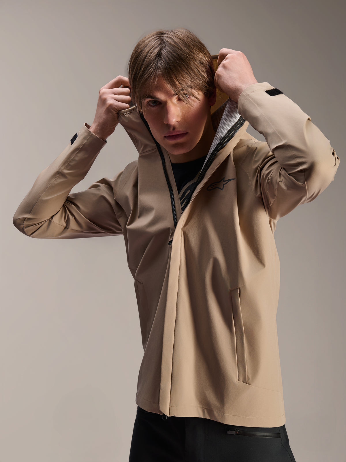 Alpinestars A-Dura Rain Jacket, MTB-cykeljakke, lys brun, model iført jakken, der trækker den elastiske hætte op, med sort brystlogo, lommer med lynlås og teknisk 2,5-lags vandtæt stof