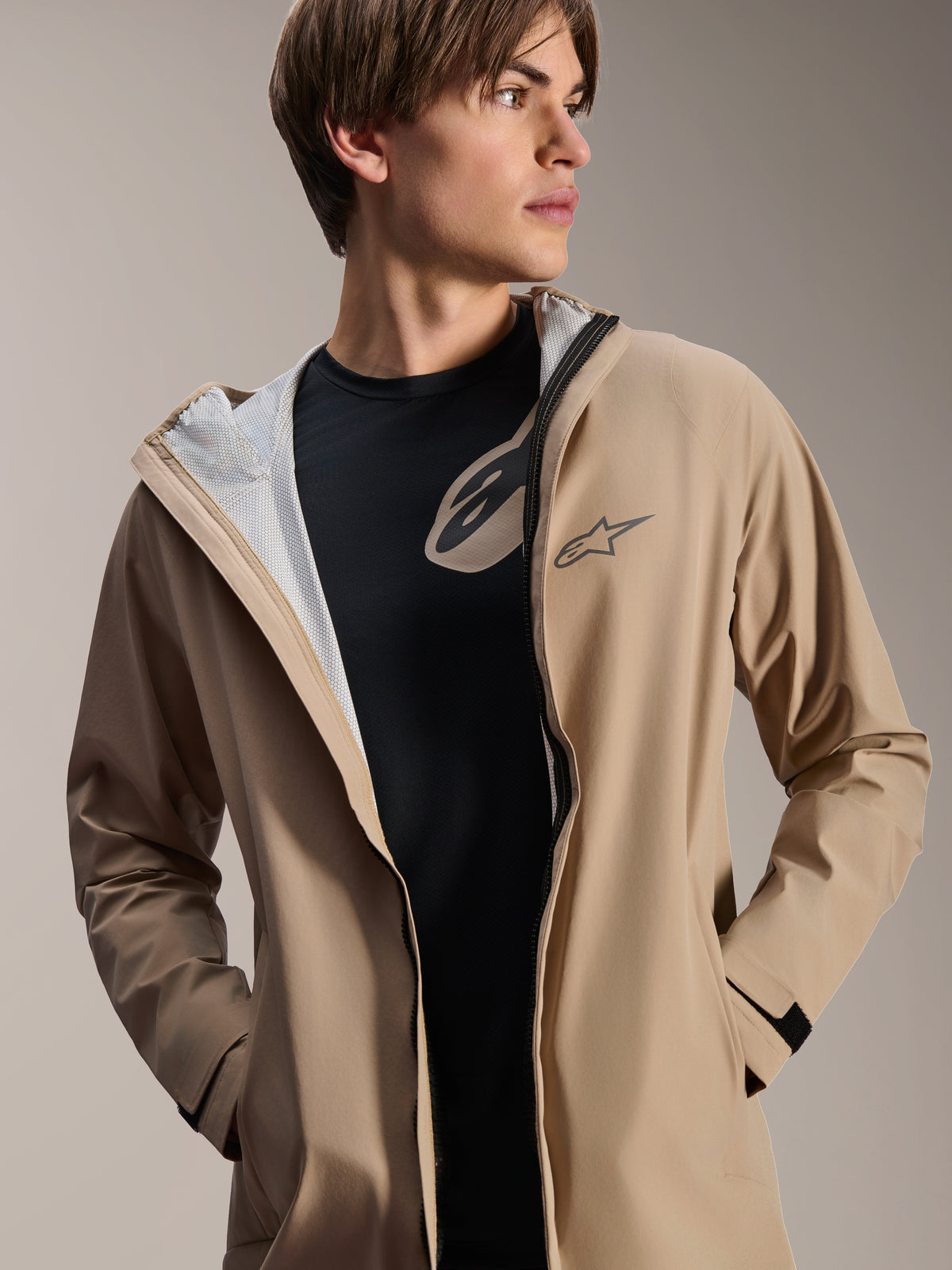 Mandlig model iført Alpinestars A-Dura regnjakke, lysebrun, lynet ned over en sort t-shirt, viser hvidt mesh-for, alsidig vejrbestandig cykeljakke