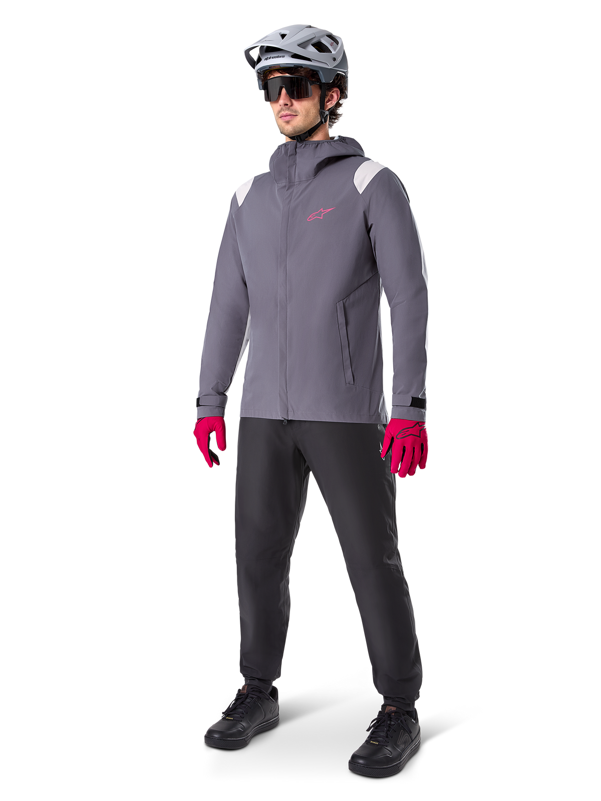 Male model wearing Alpinestars A-Dura Rain Jacket in Dark Titanium, gray hooded jacket with pink chest logo and light gray shoulder panels, paired with a gray MTB helmet, dark sunglasses, vibrant pink gloves, and black pants for off-road cycling.