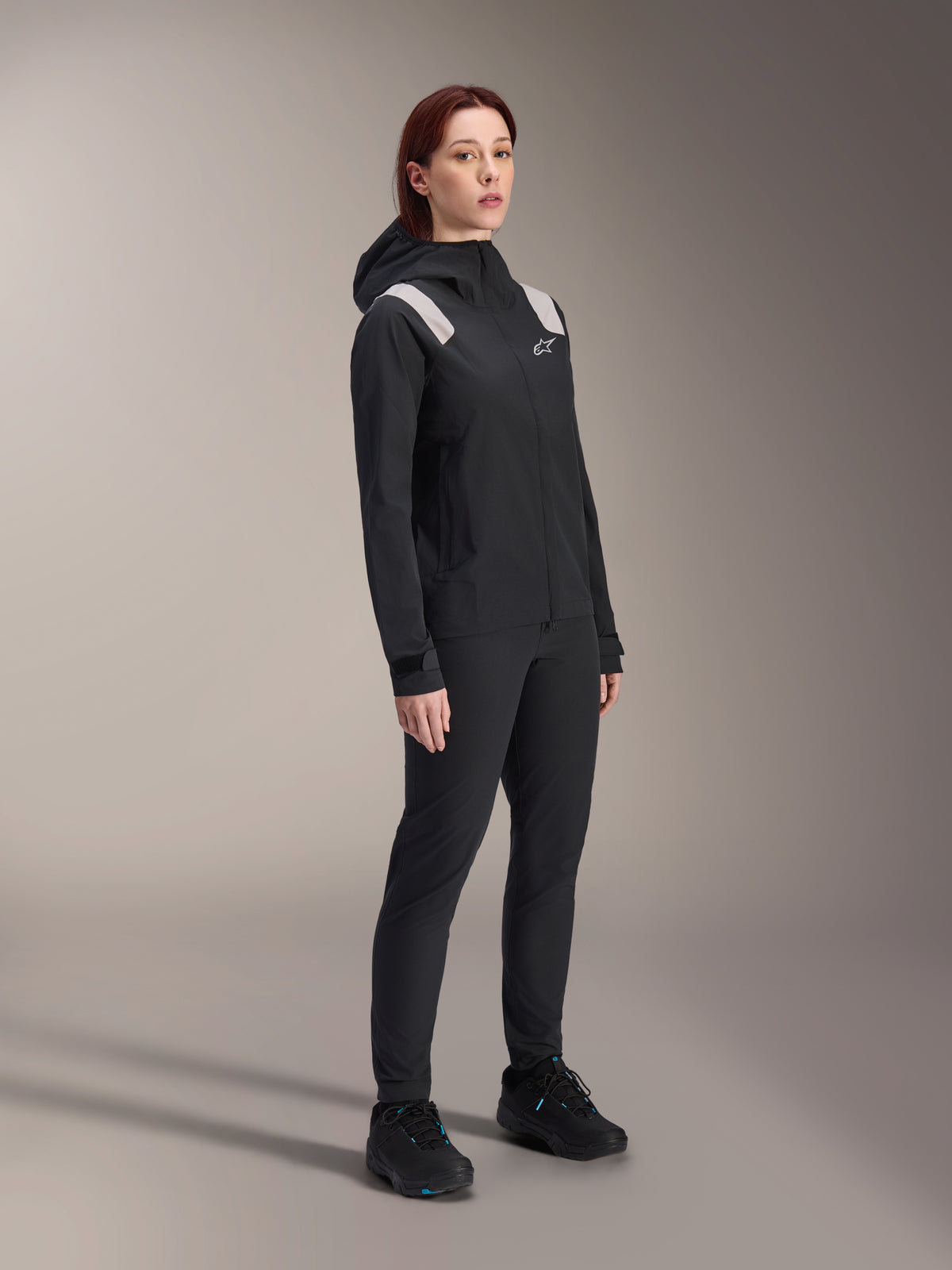 Kvinde iført Alpinestars Stella A-Dura regnjakke i sort, 2,5-lags vandtæt MTB-skal med lysegrå detaljer på skuldrene, matchende sorte bukser og cykelsko, studioportræt af performance-cykeludstyr