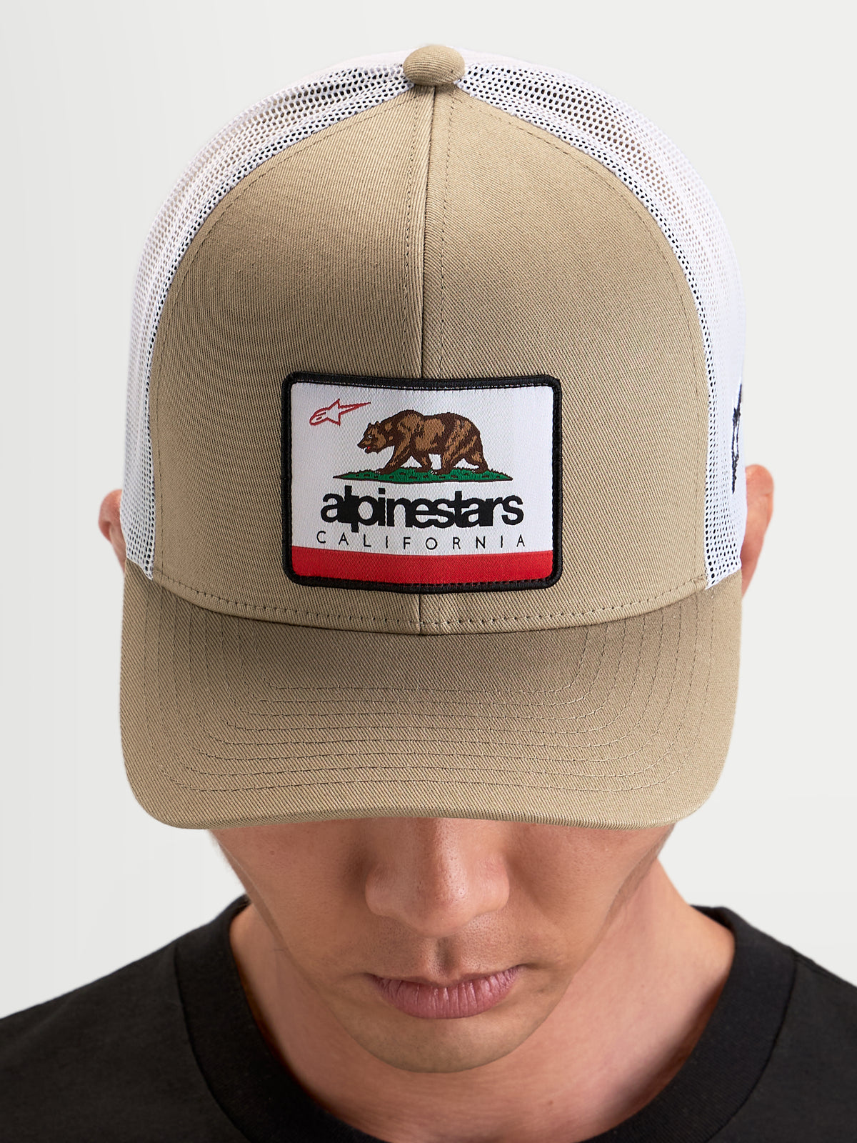 Alpinestars Cali 2.0 kasket, sportswear trucker cap i khaki og hvid med struktureret twillfront, California-bjørnepatch og buet skygge, båret af model