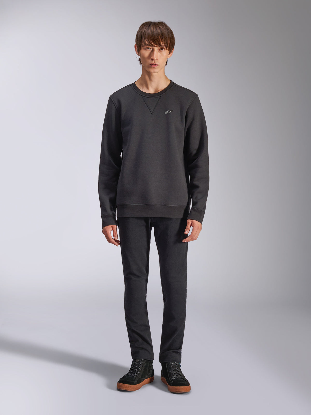 Alpinestars Positive Crew sweatshirt, afslappet sort sweatshirt med rund hals, lille hvidt logo på brystet, V-syning ved halsudskæringen, studiefoto i fuld figur med model iført sorte bukser og sneakers, motorsport-inspireret livsstilstøj