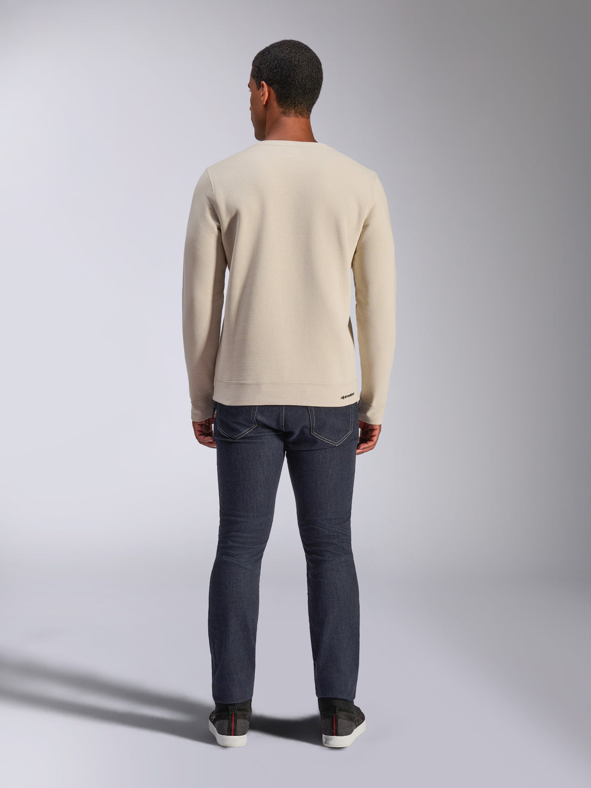 Alpinestars Positive Crew Sweatshirt, Off White, set bagfra af en mand iført en beige pullover med rund hals, ribkant ved manchetterne og forneden, kombineret med mørke jeans, afslappet motorsport-inspireret streetwear