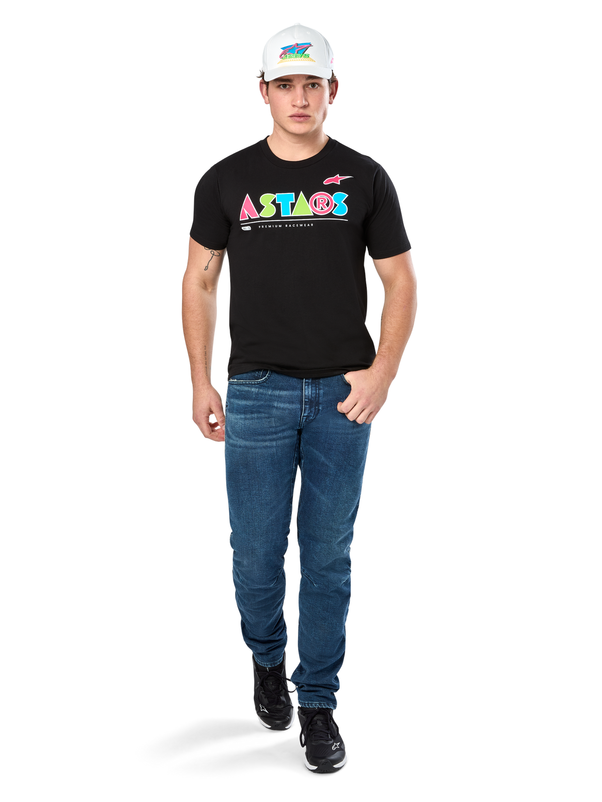 Mandlig model iført Alpinestars Cereal CSF Tee, sort kortærmet t-shirt med farverigt geometrisk ASTARS-logo, kombineret med blå jeans og hvid kasket, afslappet motorsport-inspireret streetwear.