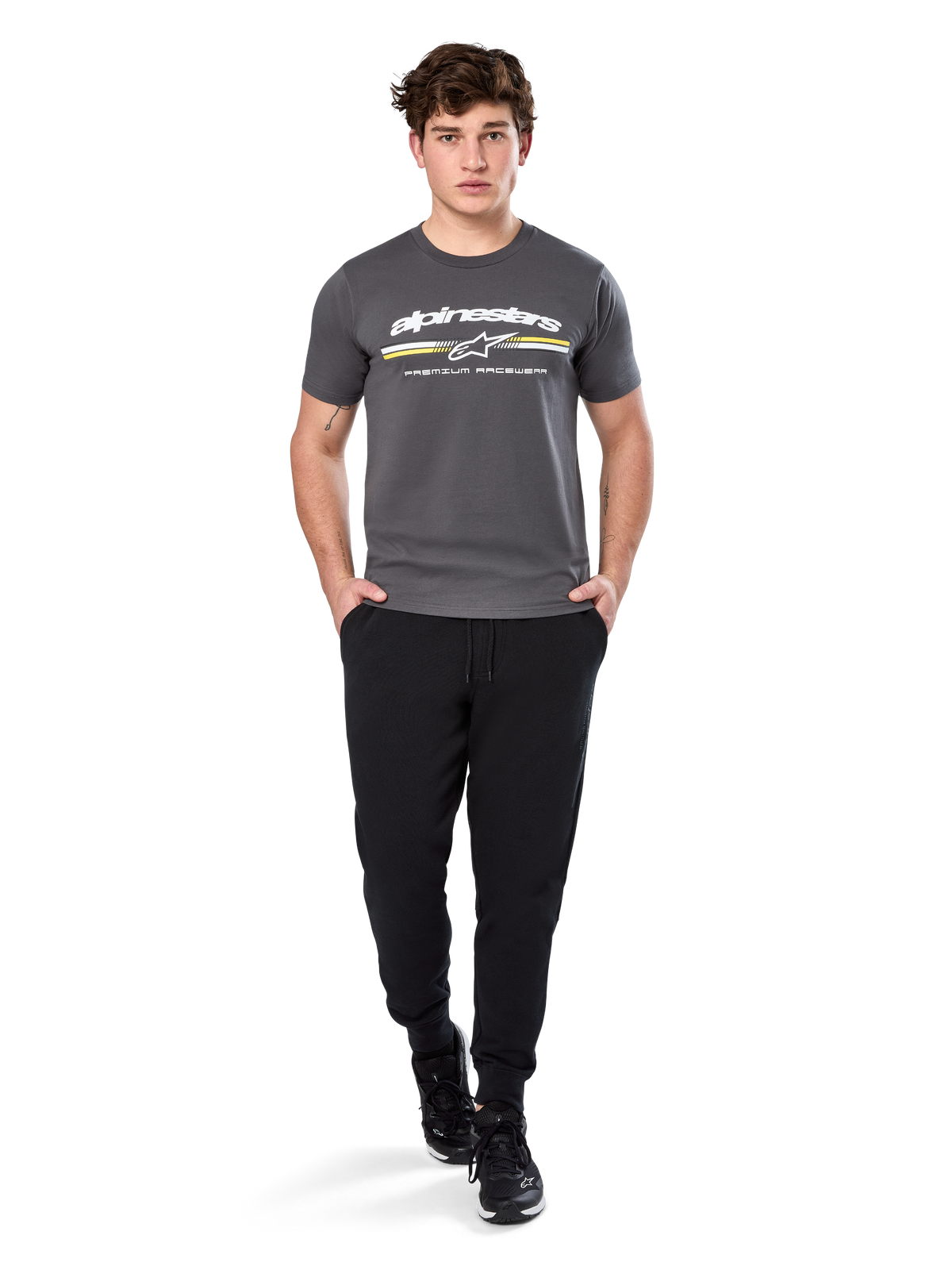 Mand iført Alpinestars Prevail Csf Tee, kortærmet T-shirt, koksgrå, hvidt Alpinestars-logo og trefarvet stribe med gule detaljer, sorte joggingbukser og sneakers, afslappet motorsport-streetwear