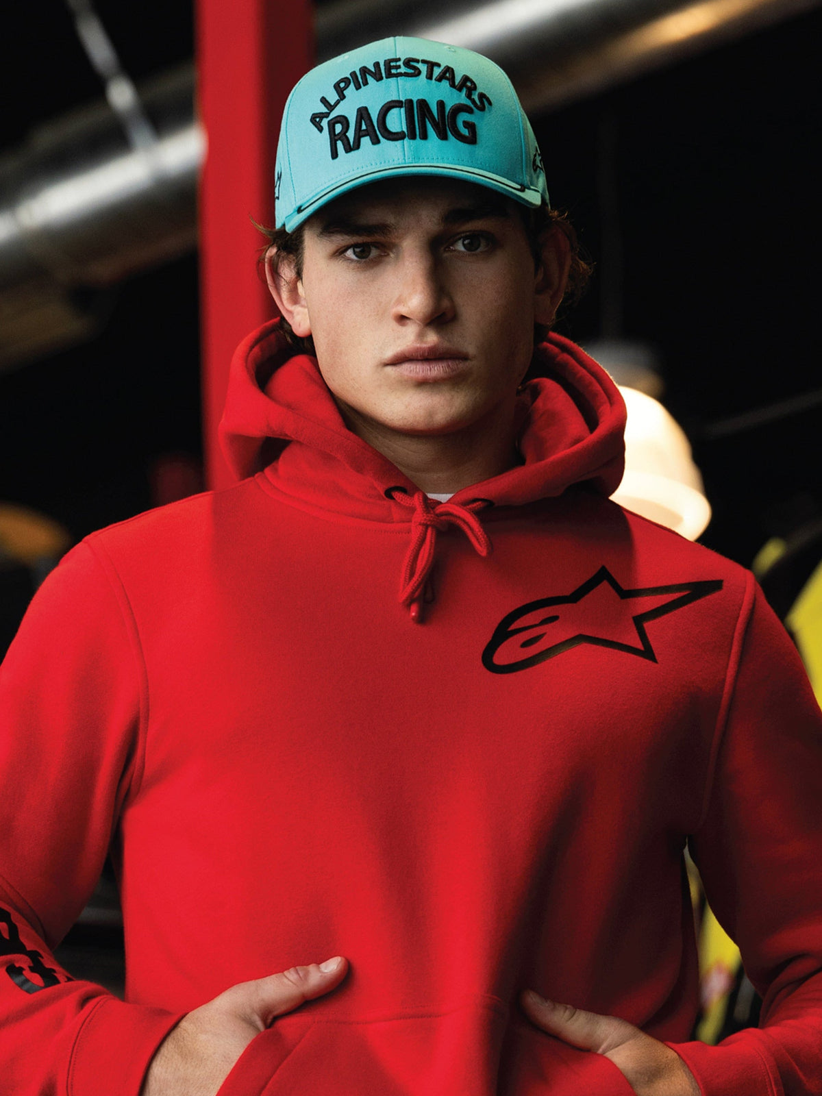 Alpinestars Luminary kasket, Aqua Blue snapback med buet skygge, sort 3D-broderi med 'ALPINESTARS RACING'-tekst, båret med en klar rød Alpinestars hoodie, afslappet motorsport lifestyle streetwear