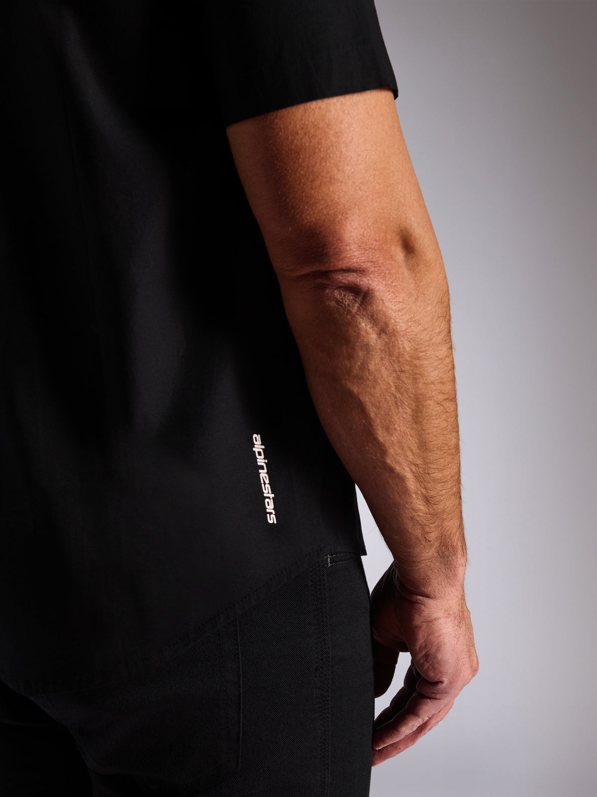 Alpinestars Strive Woven skjorte, sort kortærmet skjorte med knapper foran, lodret hvidt Alpinestars-logo på siden, nærbillede af den nederste kant og brand-logo, afslappet motorsport streetwear