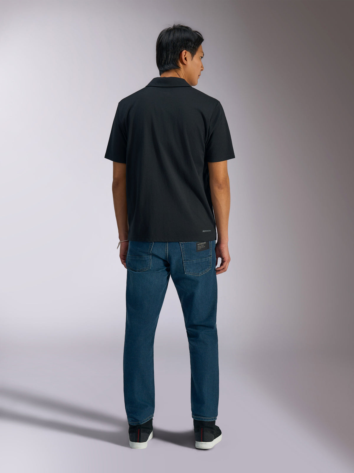 Alpinestars Impetus Sport Polo, sort, kortærmet poloshirt set bagfra med Alpinestars-branding nederst på ryggen, båret med blå jeans, afslappet motorsport-inspireret sportstøj