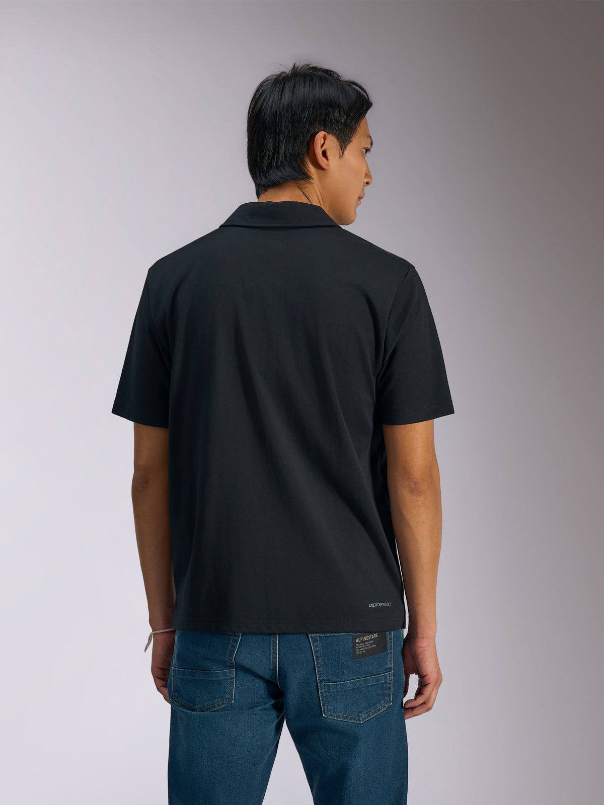 Alpinestars Impetus Sport Polo, sort kortærmet poloshirt, mand set bagfra iført polo og blå denimjeans, med lille Alpinestars-logo nederst på ryggen, afslappet motorsport-inspireret sportstøj