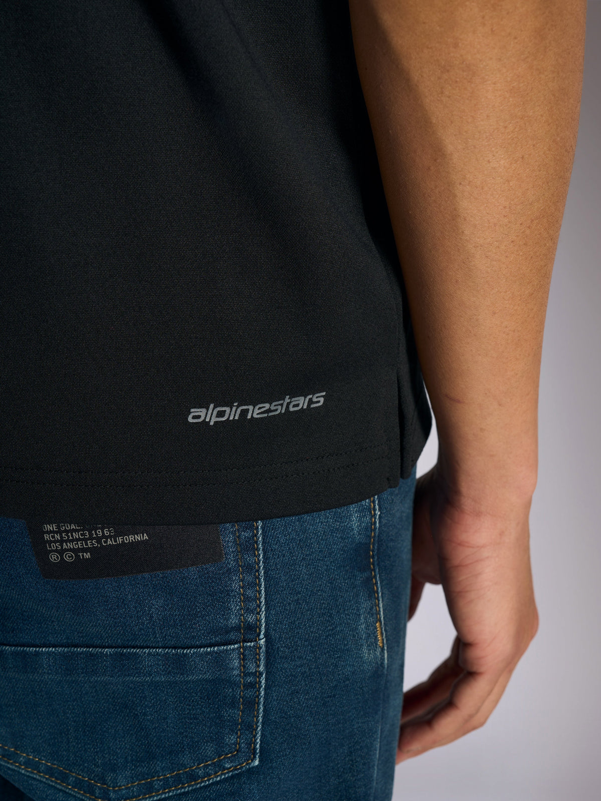Alpinestars Impetus Sport Polo, detaljeret visning af den nederste kant og sideslids, sort med gråt Alpinestars-ordmærke, afslappet motorsport-inspireret sportstøj