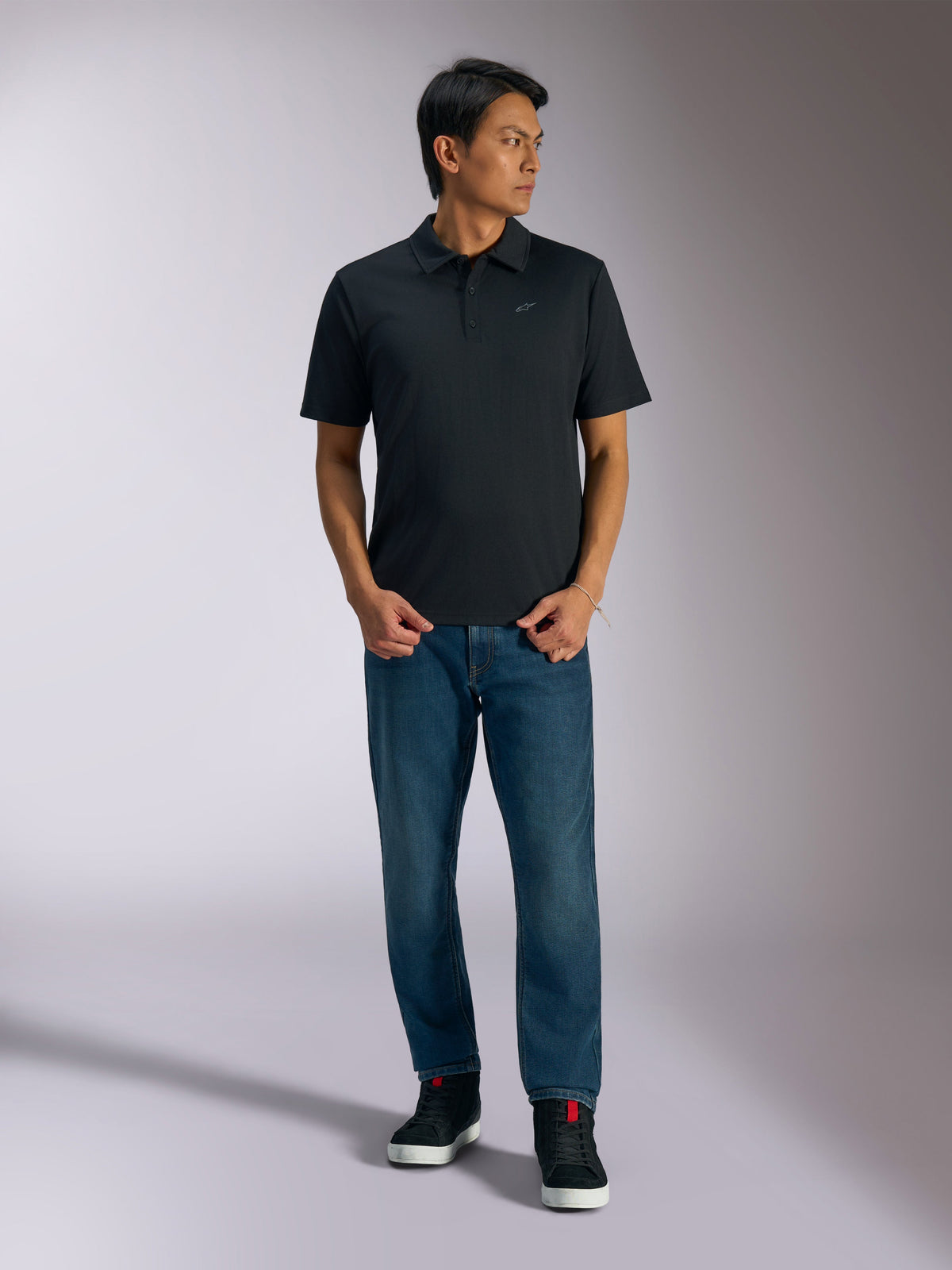 Alpinestars Impetus Sport Polo, sort kortærmet poloshirt, med tre knapper og gråt Astars-logo på brystet, båret af en mand med blå jeans og sorte high-top sneakers, afslappet motorsport-inspireret sportstøj