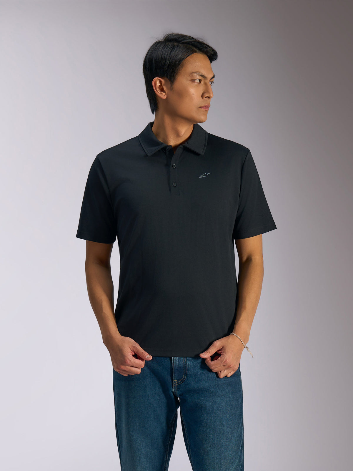 Alpinestars Impetus Sport Polo, sort kortærmet poloshirt, stolpelukning med tre knapper og selvkrave, lille gråt Astars-logo på venstre bryst, båret med blå jeans, afslappet motorsport-inspireret sportstøj