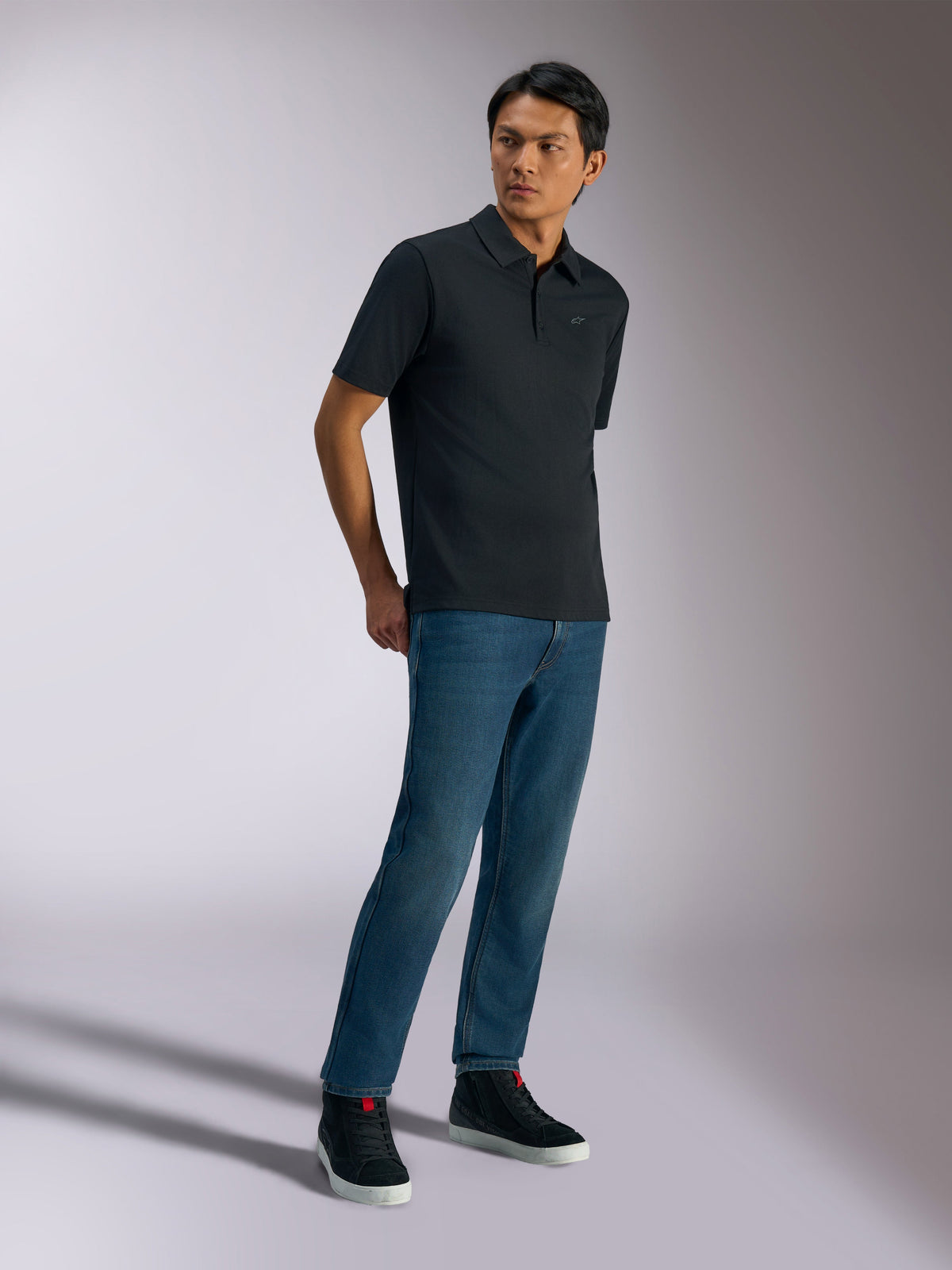 Alpinestars Impetus Sport Polo, kortærmet poloshirt, sort, med et diskret brystlogo og stolpelukning med tre knapper, båret af en model med blå jeans og sorte sneakers, afslappet motorsport-inspireret livsstilstøj