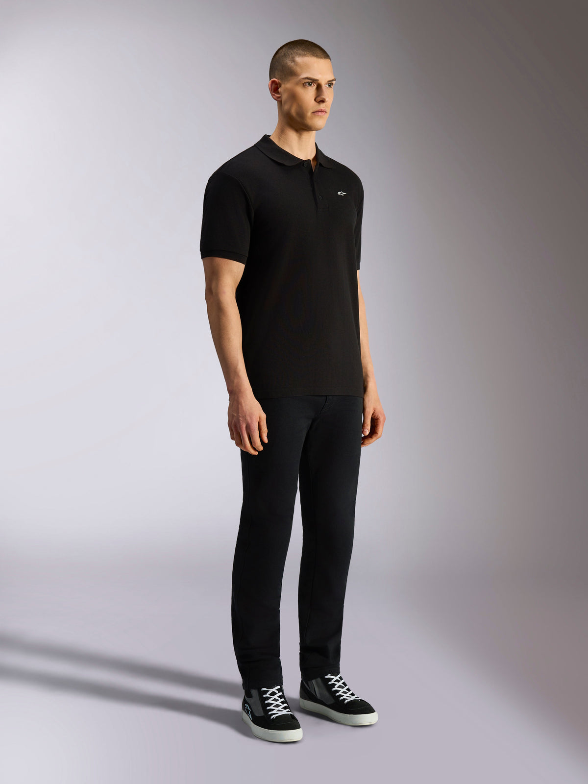 Alpinestars Endeavor Classic Polo, sort kortærmet poloshirt, piqué-strik med tre-knaps stolpelukning, lille hvidt Astars-logo på venstre bryst, båret med sorte bukser og sneakers, afslappet motorsport-streetwear