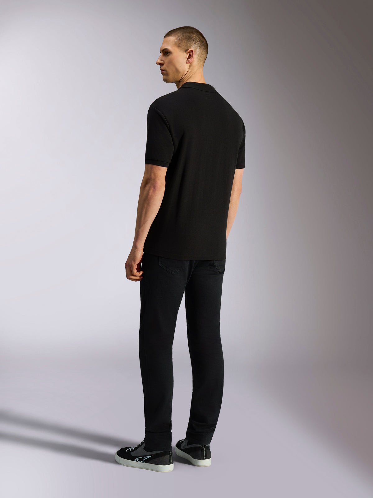 Alpinestars Endeavor Classic Polo, kortærmet piqué-poloshirt, sort, set bagfra på mandlig model iført sorte bukser og sneakers, afslappet motorsport-inspireret streetwear