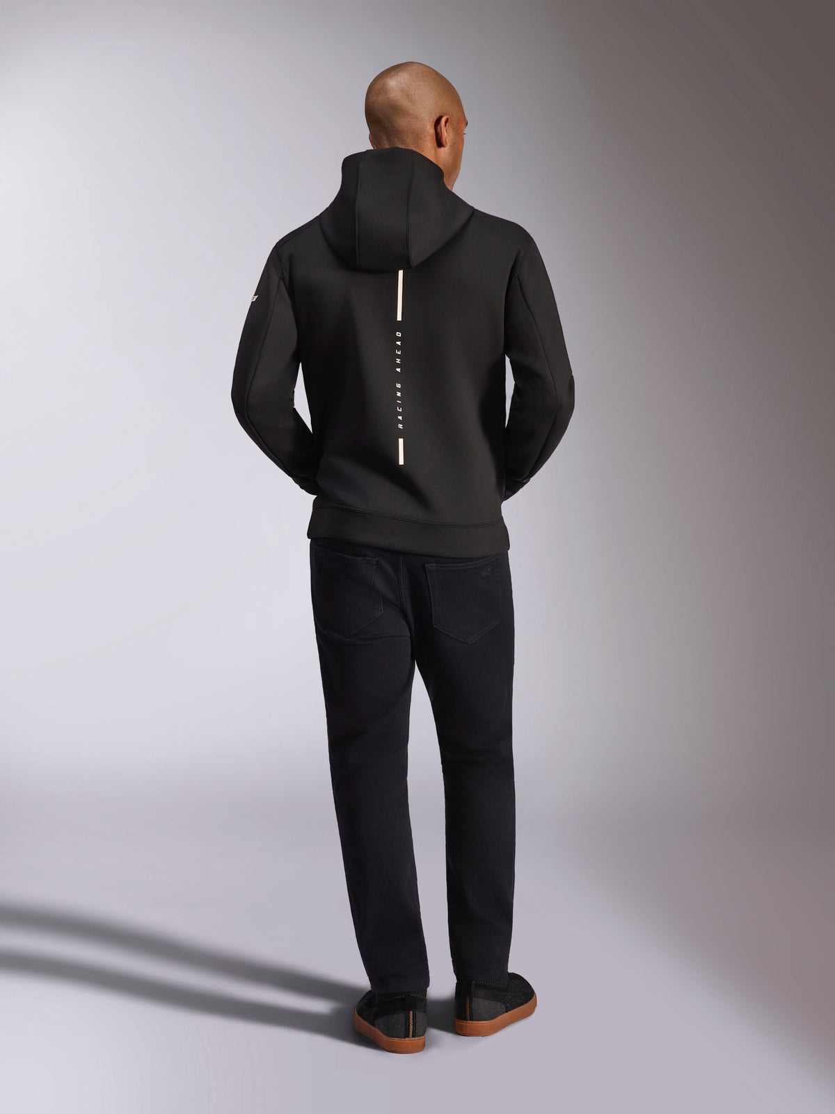 Alpinestars Insight Hoodie, sort, set bagfra på model, med et lodret hvidt stribelogo på ryggen med teksten "Racing Ahead", afslappet sportstøj-hættetrøje i tri-blend dobbeltlags fleece, kombineret med sorte bukser.