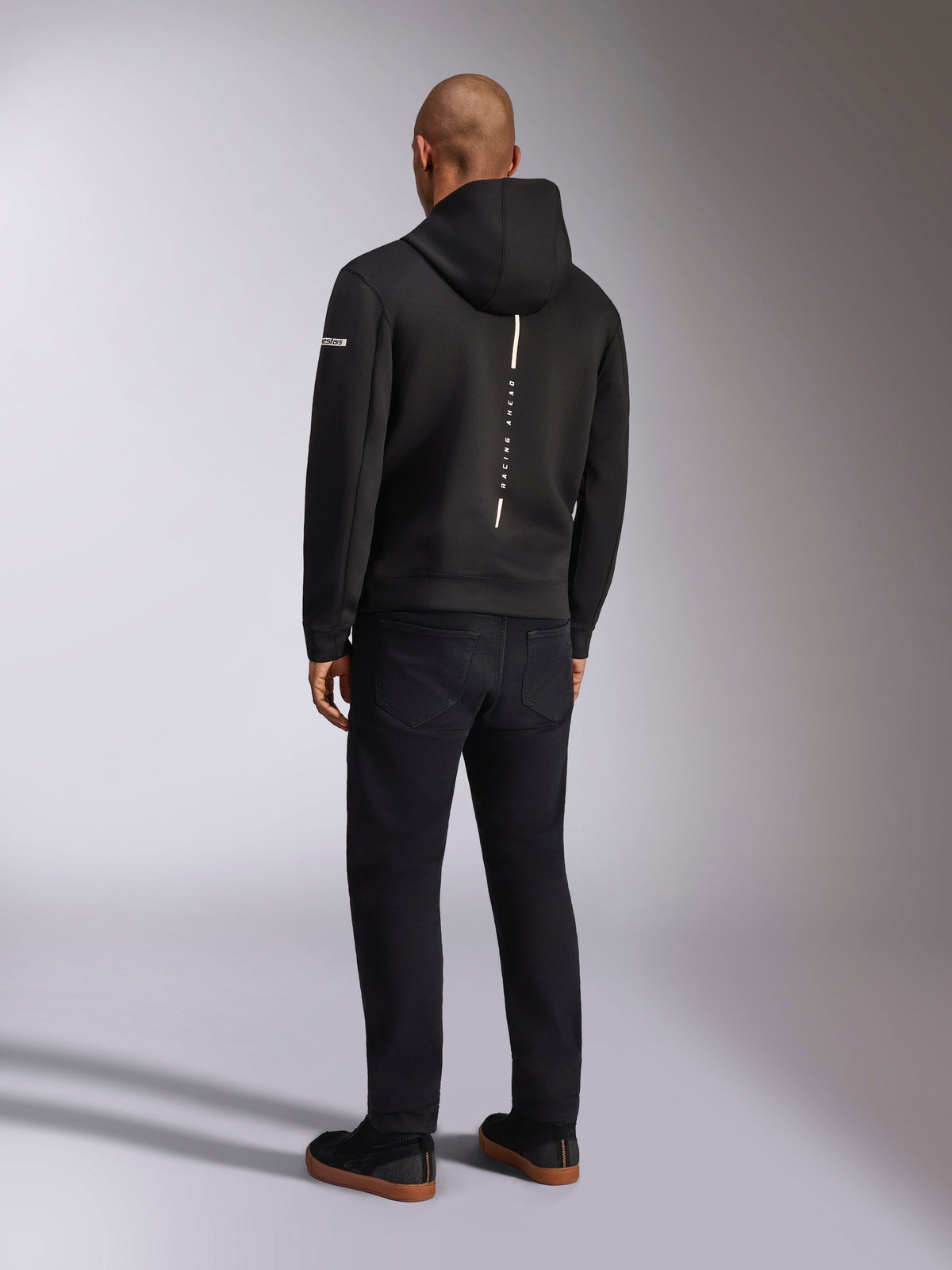 Alpinestars Insight Hættetrøje, sort sportshættetrøje, set bagfra med en lodret hvid stribe med 'RACING AHEAD' branding, logo på ærmet, kombineret med sorte bukser og sneakers, afslappet motorsport-inspireret design