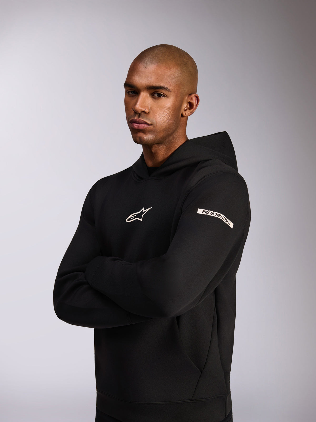 Alpinestars Insight Hoodie, afslappet sportstøj-hættetrøje, sort, hvidt Astars-logo på brystet og tekstlogo på overarmen, model iført hættetrøje med armene over kors, førsteklasses dobbeltsidet fleece-konstruktion