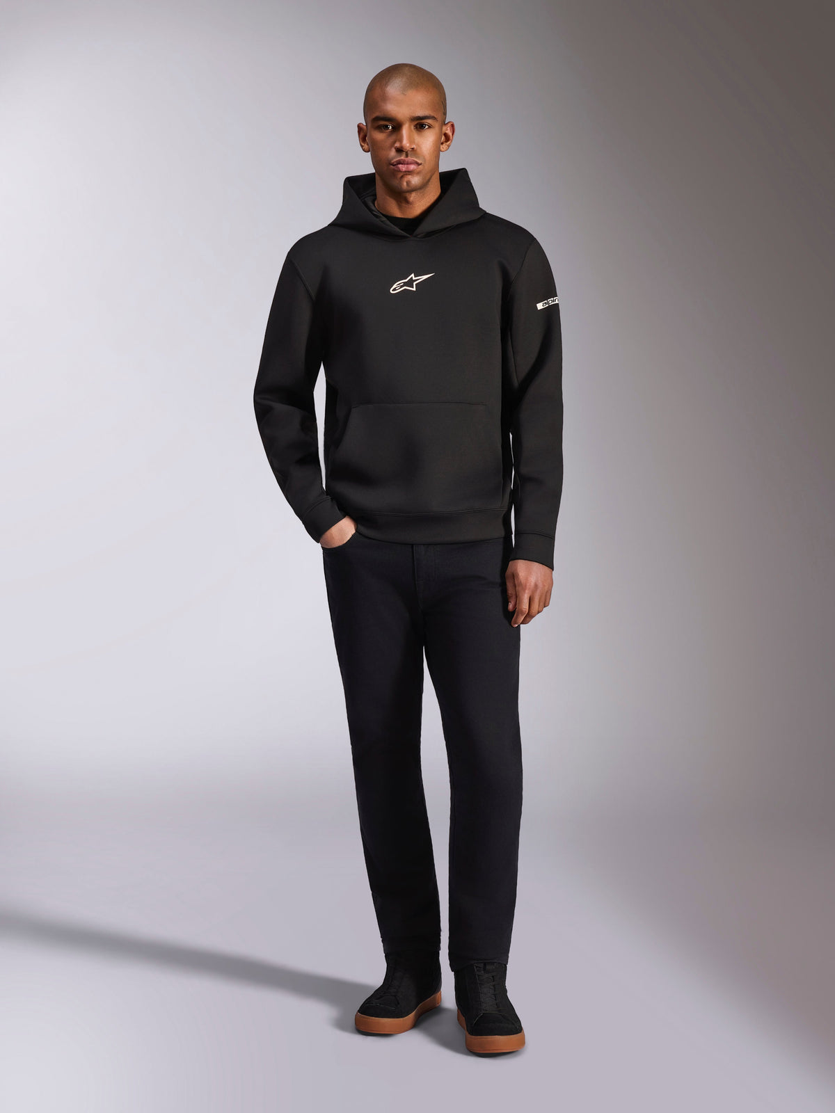 Alpinestars Insight hættetrøje, sort pullover-hættetrøje med hvidt Astars-logo på brystet og ærmet, båret af en model med sorte bukser og sorte sneakers, afslappet motorsport-inspireret streetwear