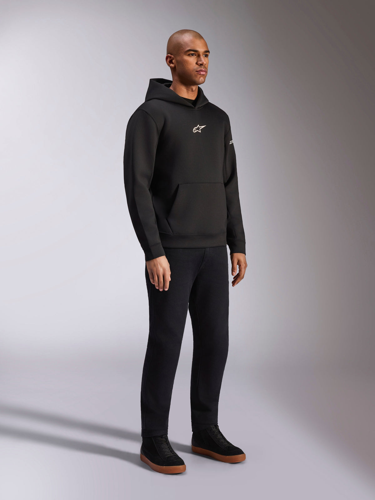 Alpinestars Insight Hættetrøje, sort, afslappet pullover-fleecehættetrøje med hvidt Astars-logo på brystet, kængurulomme, kombineret med sorte bukser og sneakers, studiebillede i fuld figur, motorsport-inspireret streetwear