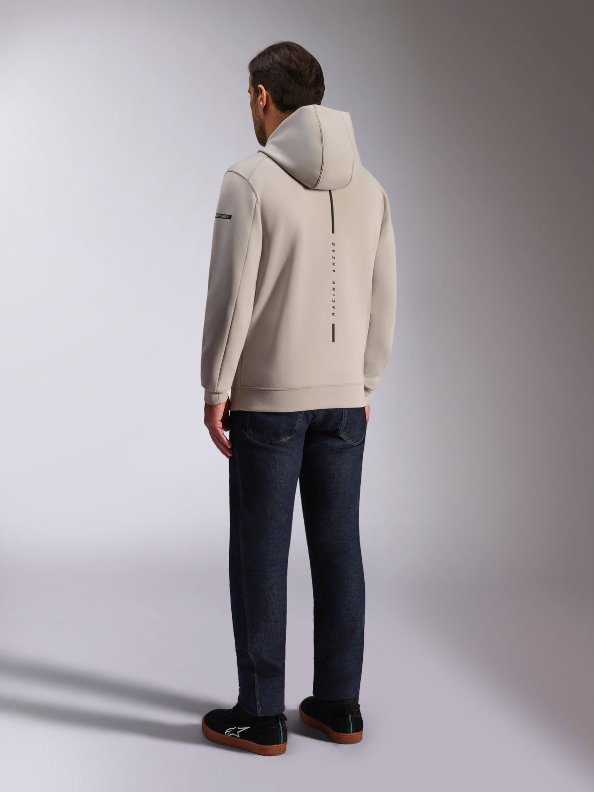 Alpinestars Insight hættetrøje, set bagfra i cementgrå, lodret sort stribe med 'RACING AHEAD'-tekst, afslappet streetwear-design, båret med mørke jeans og sorte sko.