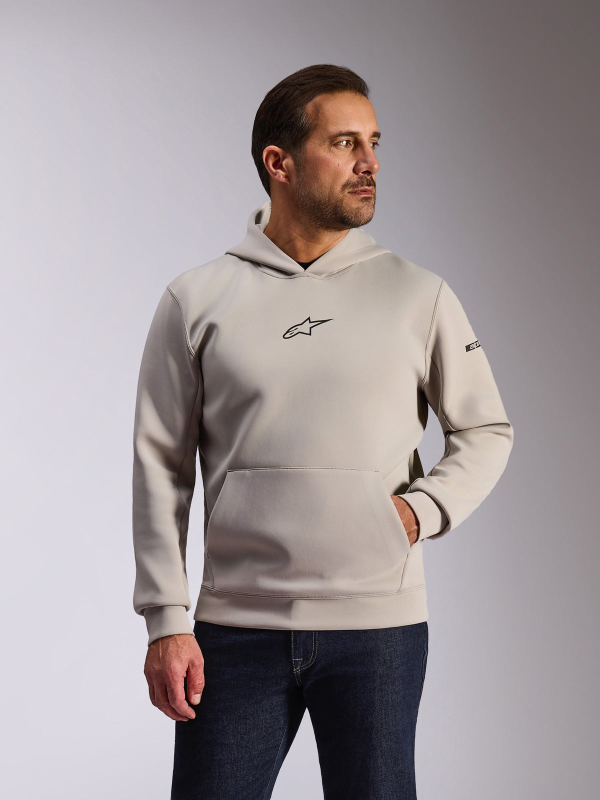 Alpinestars Insight hættetrøje, afslappet sportstøj, cementgrå, pullover-design med kængurulomme og lille sort Astars-logo på brystet, lange ærmer med Alpinestars-tekstlogo på venstre arm, båret af en model med mørke denimjeans, livsstils-motorsportstøj