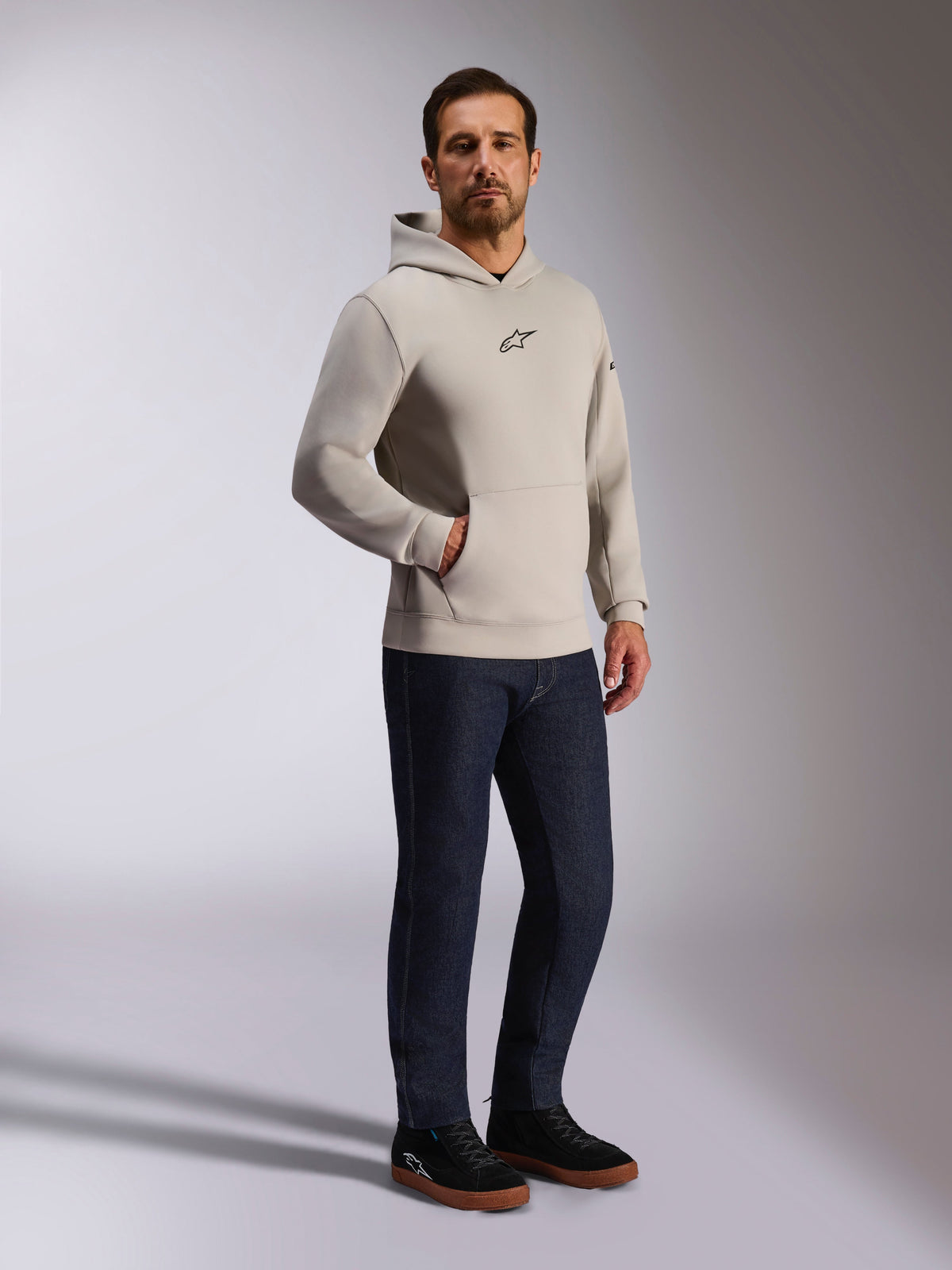 Alpinestars Insight Hoodie, afslappet streetwear, cementgrå med sort Astars-logo på brystet, pullover-design med kængurulomme, båret med mørkeblå jeans og sorte sneakers, moderne motorsport-inspireret stil