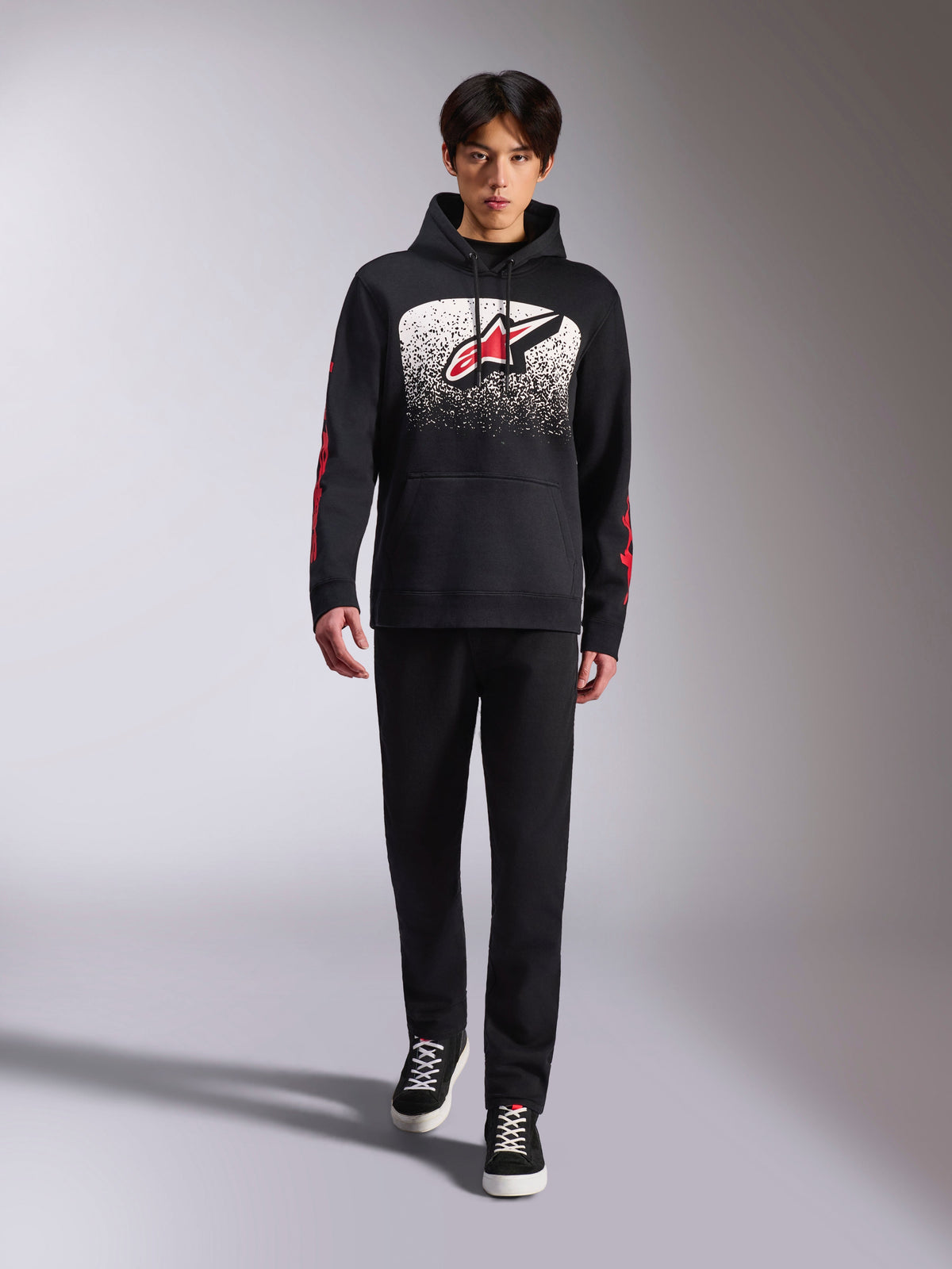 Alpinestars Obsessed hættetrøje, sort pullover-hættetrøje, stort hvidt og rødt Astars-logo med MX-graduering, røde word-logoer på ærmerne, båret af model i et helt sort afslappet outfit, motorsport-inspireret livsstilstøj