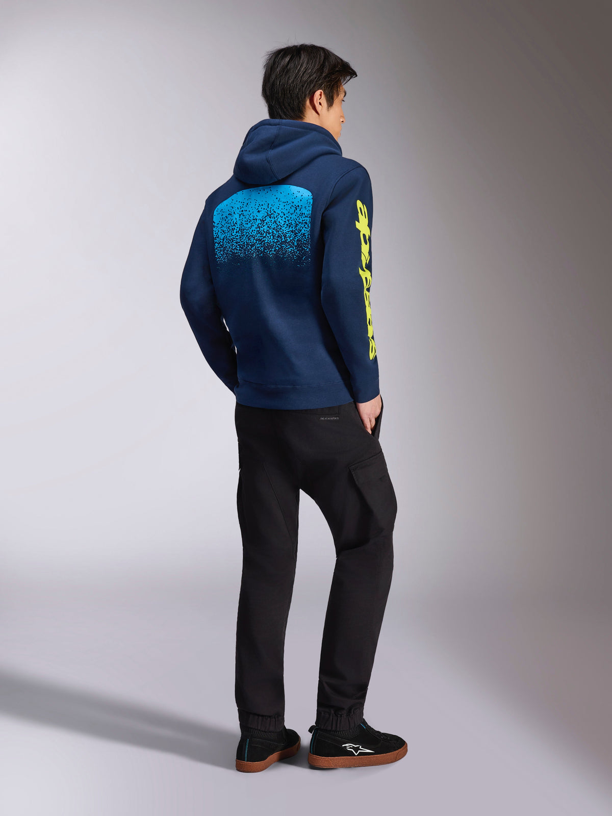 Alpinestars Obsessed hættetrøje, marineblå afslappet sweatshirt, set bagfra viser lyseblå MX-gradueringsgrafik og gult Alpinestars-logo på ærmet, båret med sorte bukser og sneakers