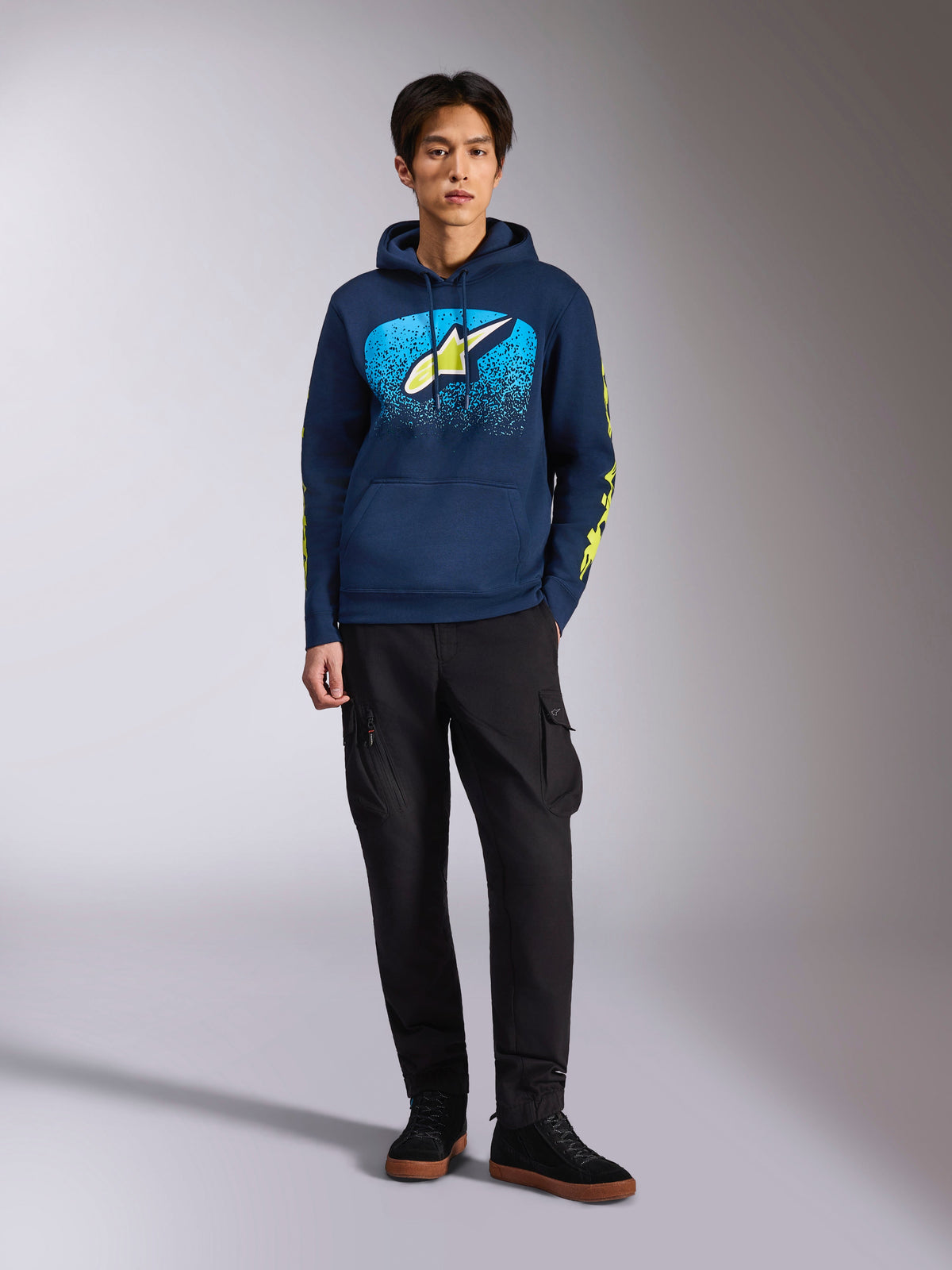 Alpinestars Obsessed hættetrøje, mørkeblå pullover-hættetrøje, Astars-logo på fronten med blå MX-graduering og limegul stjerne, limegule Alpinestars-logoer på ærmerne, båret af en model med sorte cargobukser og sorte sneakers, afslappet motorsport-inspireret streetwear
