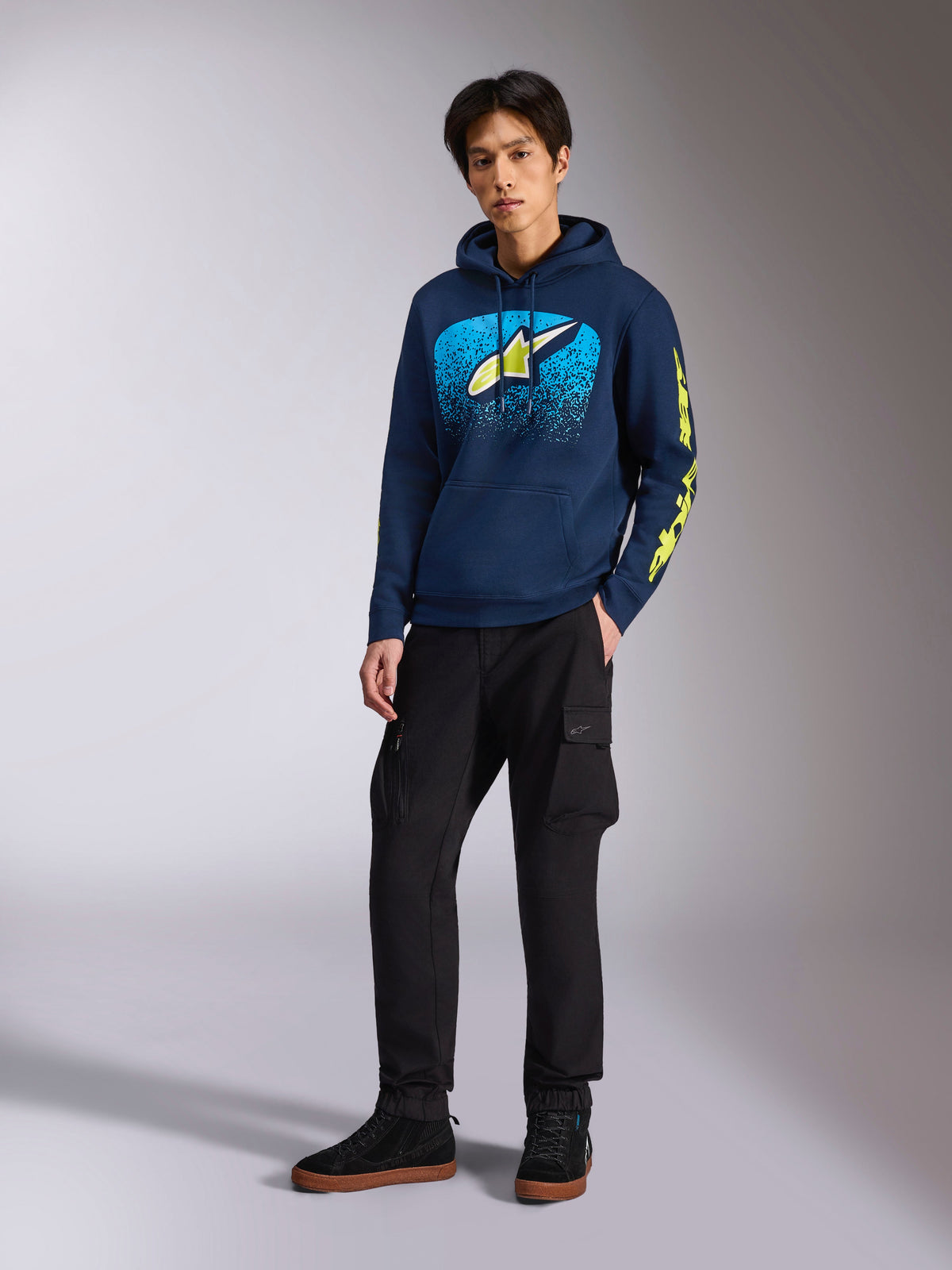 Alpinestars Obsessed Hoodie i navy, afslappet pullover-fleece med lyseblå og limegul grafik på fronten, limegult Alpinestars-logo på ærmet, båret af en model med sorte cargobukser og sorte sneakers, motorsport-inspireret streetwear