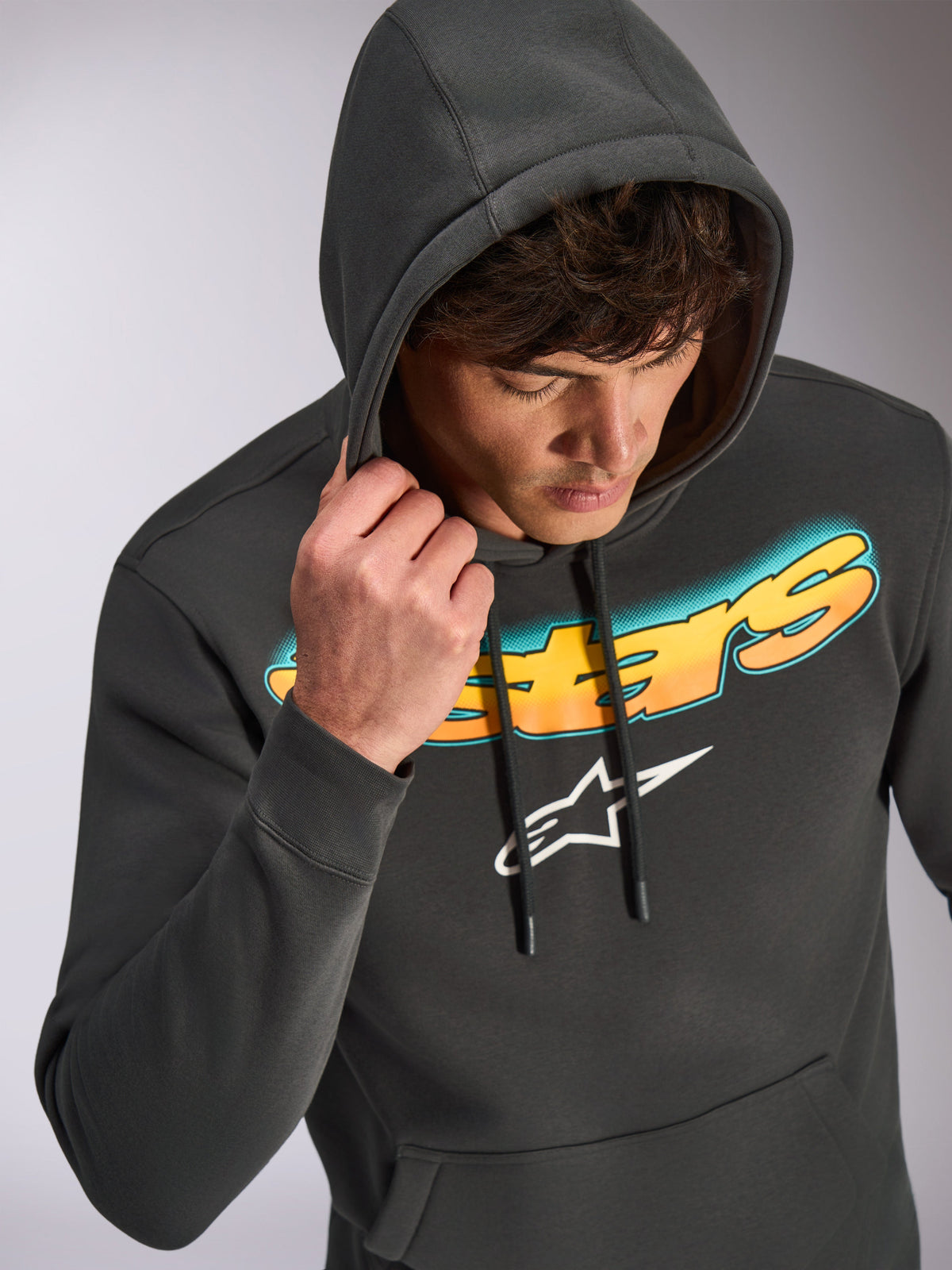 Alpinestars Ellipsoid hættetrøje, gunmetal grå, afslappet pullover-hættetrøje med Astars-tekst i en farveovergang fra cyan til orange og et hvidt logo, båret af model med hætten oppe, motorsportsinspireret streetwear