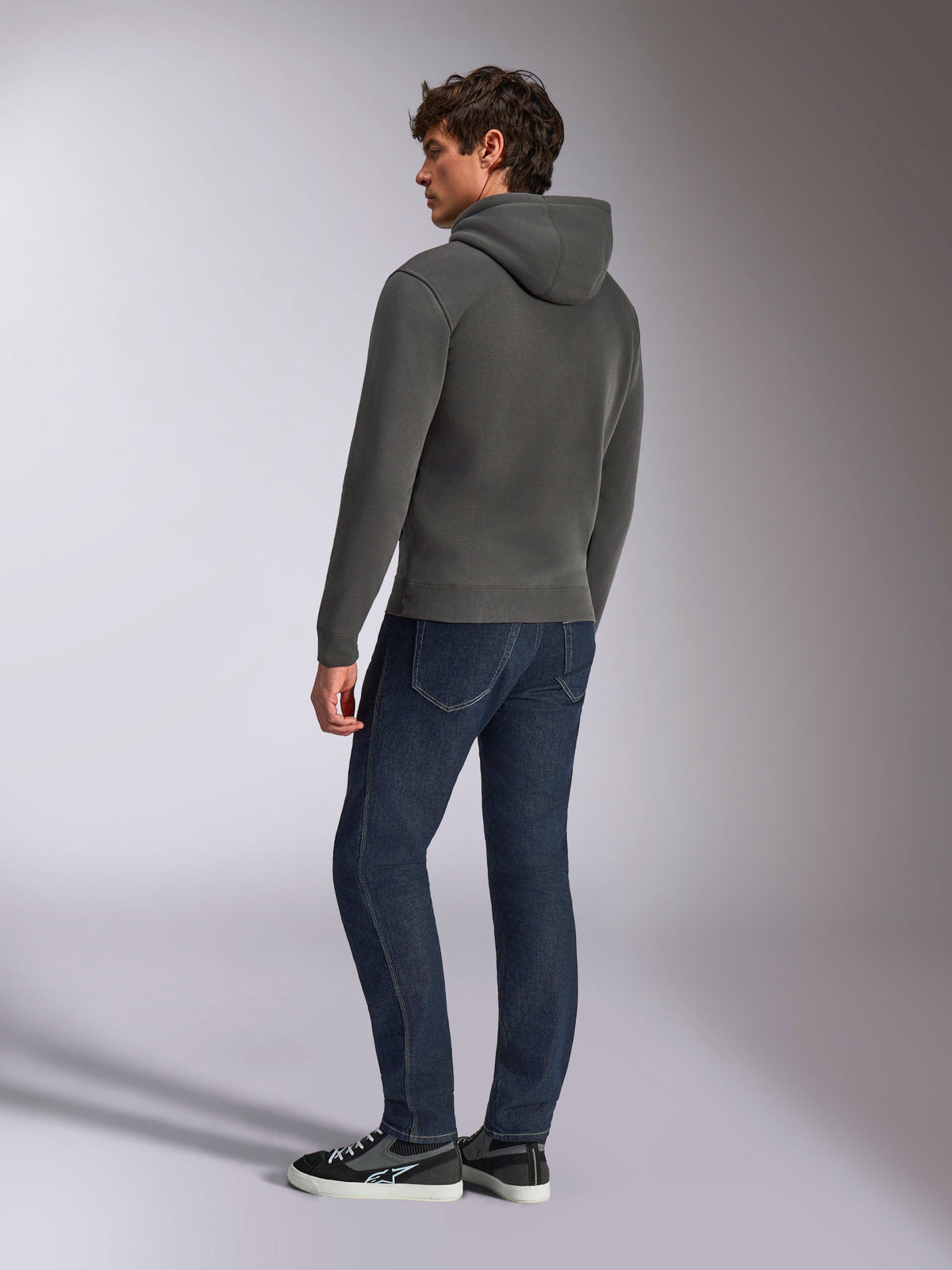 Alpinestars Ellipsoid hættetrøje, afslappet sportstøj, gunmetal-grå, set bagfra på mandlig model, hætte med snøre, lange ærmer, ribstrikkede manchetter og kant forneden, kombineret med mørke jeans og sneakers