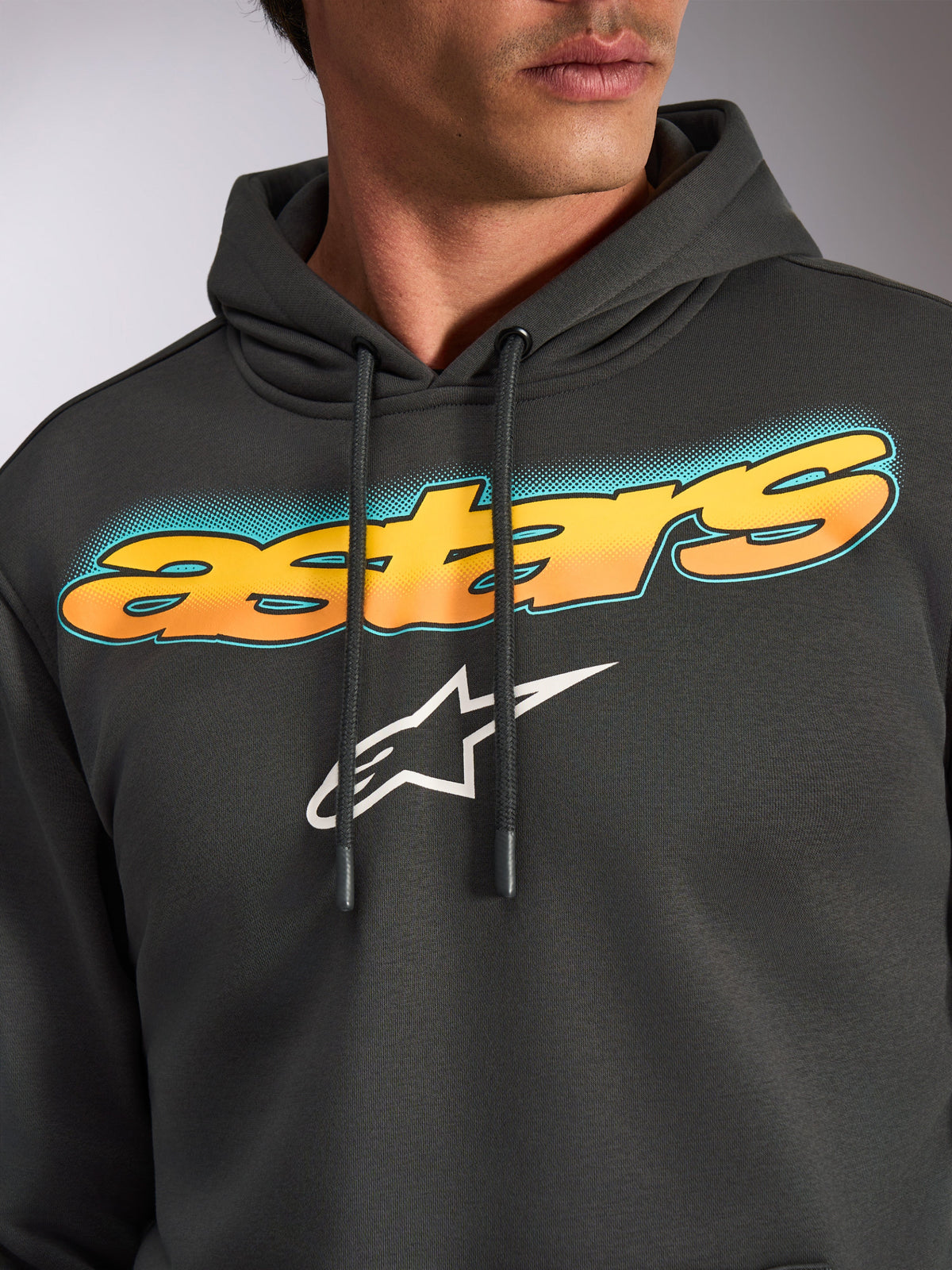 Alpinestars Ellipsoid hættetrøje, Gunmetal grå, nærbillede af brystet med buet 'astars'-grafik med gul-til-orange farveovergang og teal-accenter, hvidt Alpinestars stjerne-logo, afslappet motorsport-inspireret streetwear-fleece