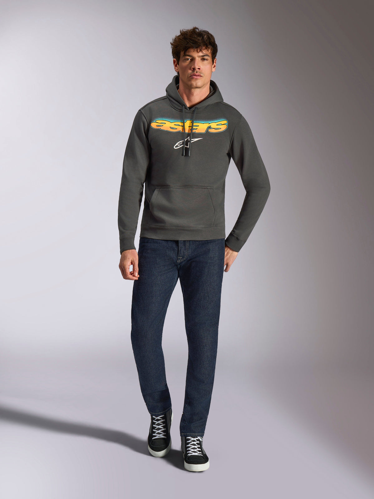 Alpinestars Ellipsoid hættetrøje, afslappet sportstøj, gunmetal grå med orange og gult Astars-logo med farveovergang og hvidt symbol, hætte med snoretræk, båret af en mandlig model med mørkeblå jeans og sorte sneakers, moderne motorsport-inspireret streetwear