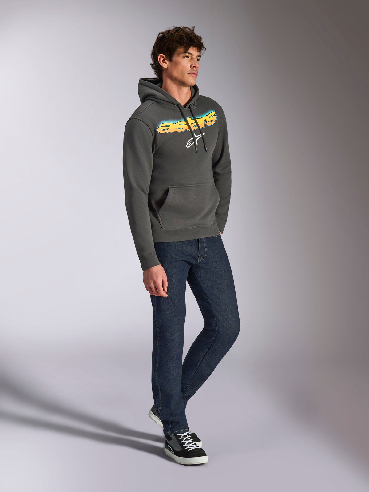 Alpinestars Ellipsoid hættetrøje, gunmetal grå pullover-fleece, buet Astars-tekst på fronten med blå og gul farveovergang og hvidt logo, stylet med mørke jeans og sorte sneakers, afslappet streetwear.