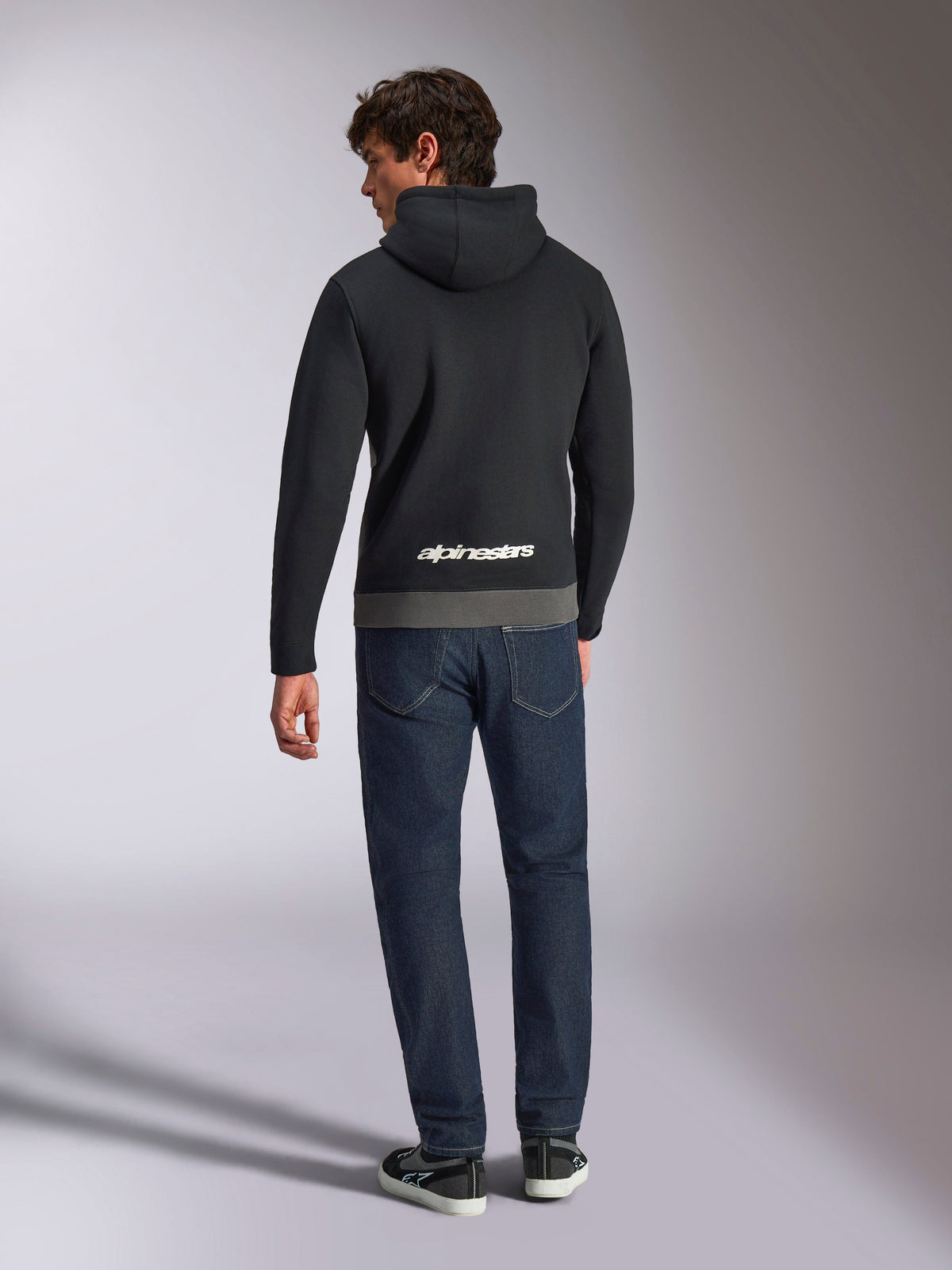 Alpinestars Parallel hættetrøje, mand set bagfra iført sort og gunmetal farveblokket pullover-fleece, hvidt Alpinestars ordlogo på den nederste del af ryggen, mørkeblå jeans, afslappet motorsport-inspireret streetwear