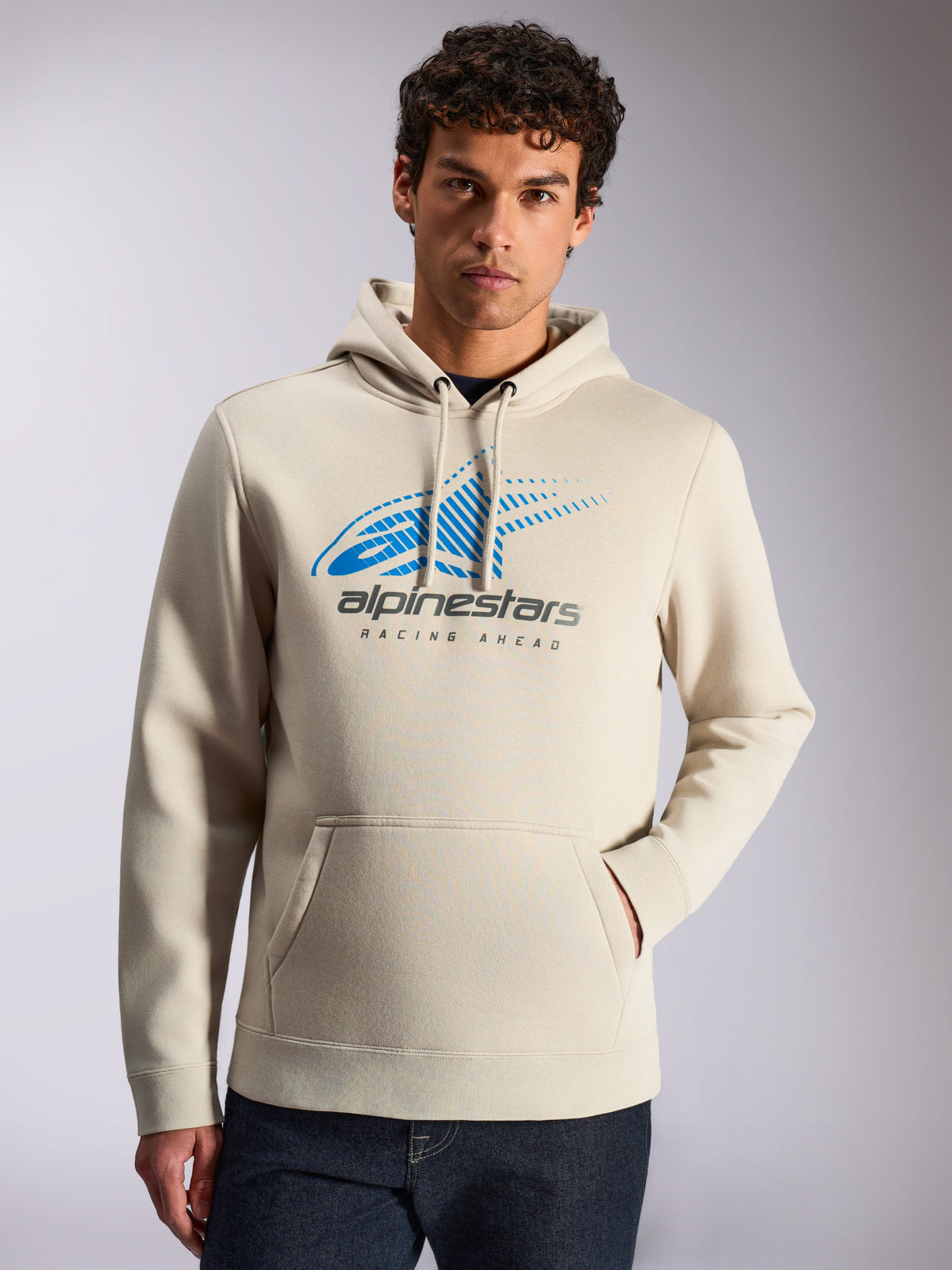 Alpinestars Pulse Hoodie, afslappet streetwear, sandfarvet, blåt og gråt Astars Combo-logo på fronten med speed line fade og 'Racing Ahead'-tekst, hætte med snoretræk, kængurulomme, båret af en model med mørke jeans