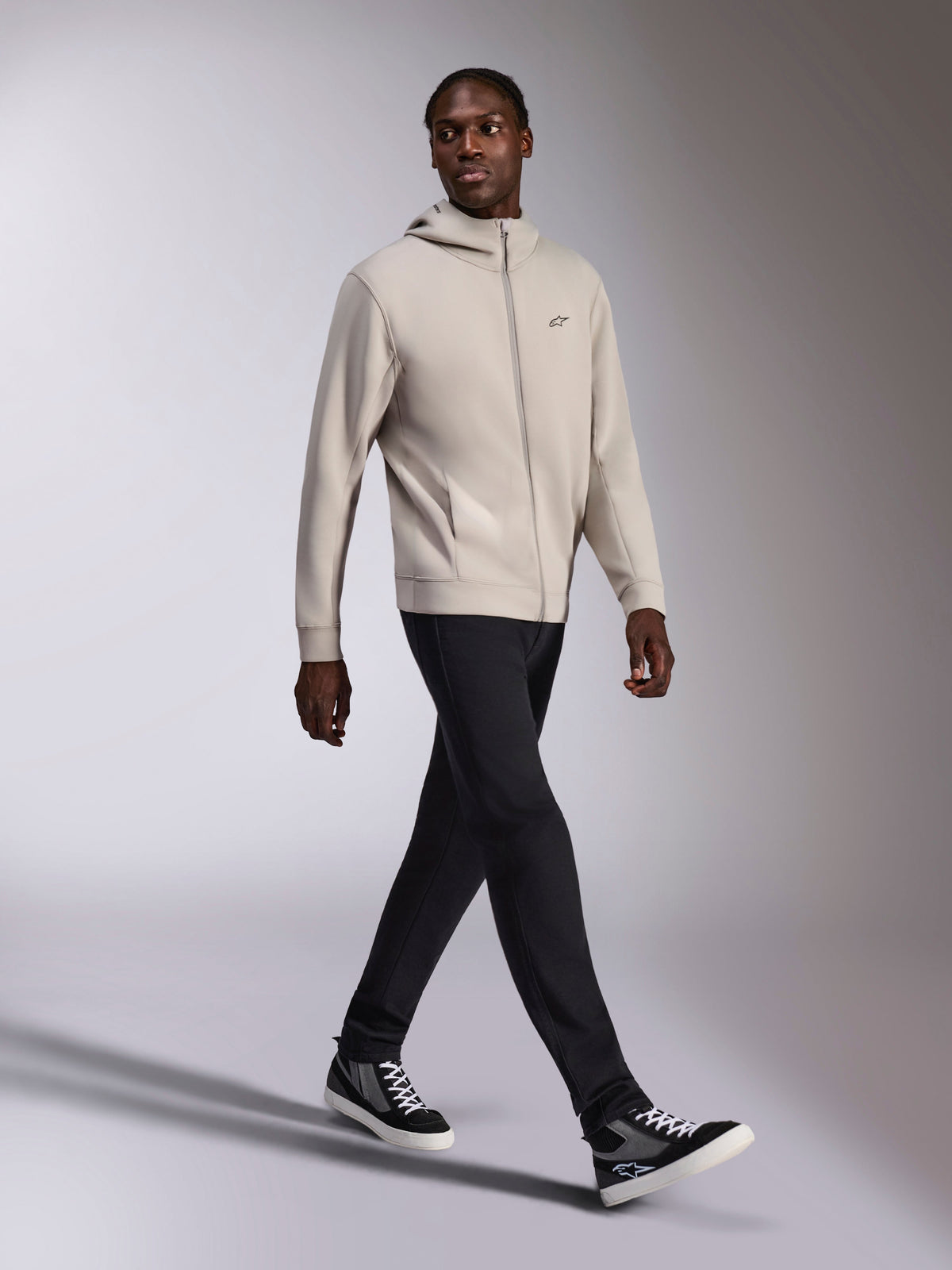 Alpinestars Cypher Hoodie, cementgrå fleece med gennemgående lynlås, højhalset hætte, lille Astars-logo på brystet, båret af en gående model, kombineret med sorte bukser og sort-grå sneakers, afslappet motorsport-inspireret sportstøj