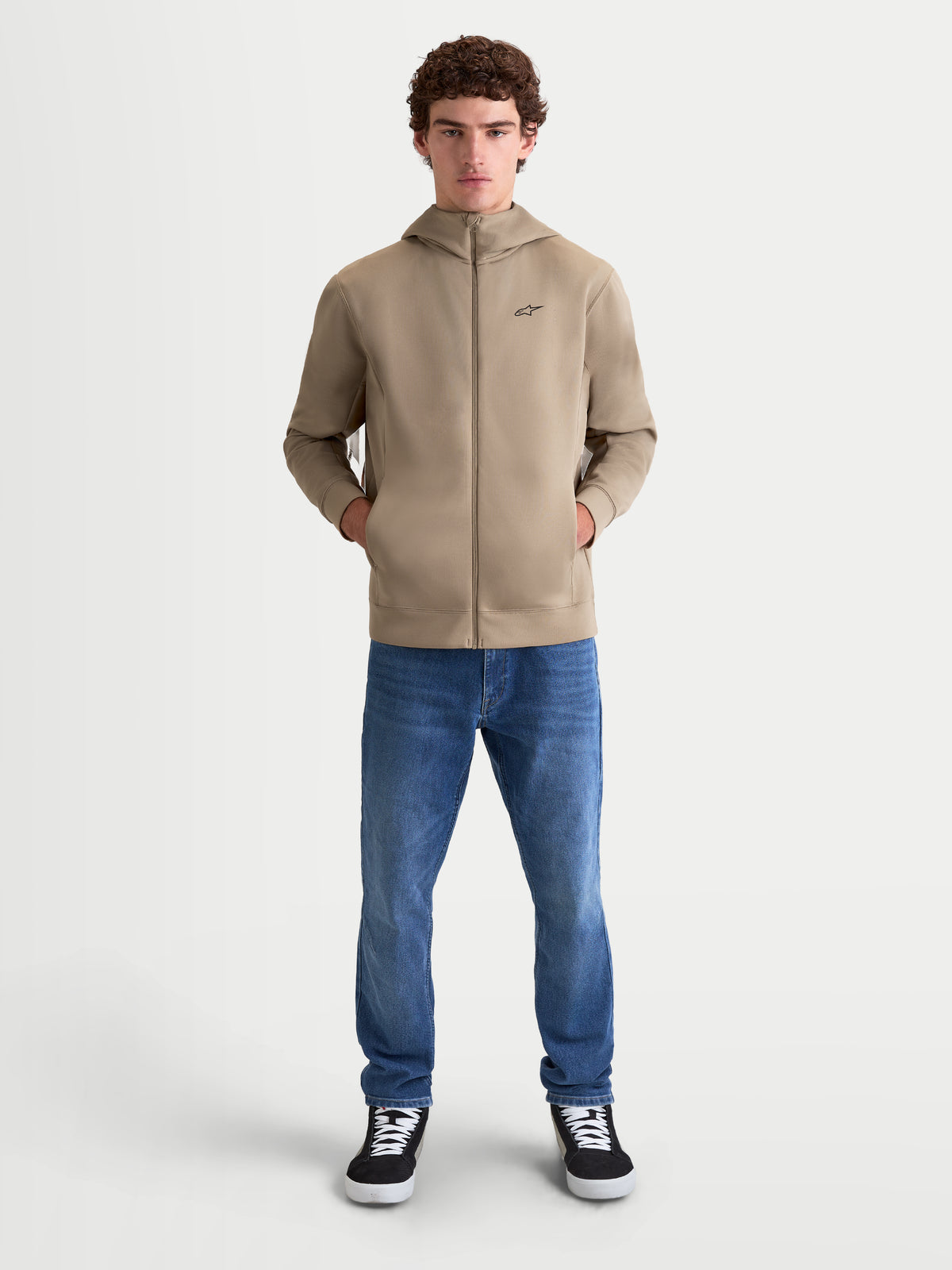 Alpinestars Cypher hættetrøje, herre-sportswear fleece med fuld lynlås i putty beige, højhalset hætte, diskrete lommer i sømmen, model iført beige hættetrøje og blå jeans, motorsport-inspireret design med et lille sort logo på brystet.