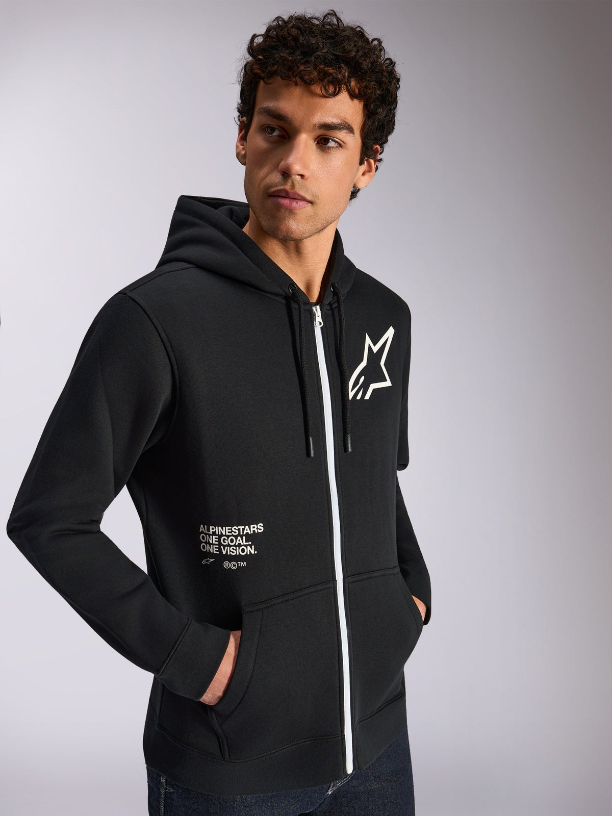 Alpinestars Transverse hættetrøje, afslappet sportstøj, sort med hvid lynlåsdetalje, hætte med snoretræk, Alpinestars stjerne-logo på brystet, 'One Goal One Vision'-grafik, båret af mandlig model med mørkt krøllet hår