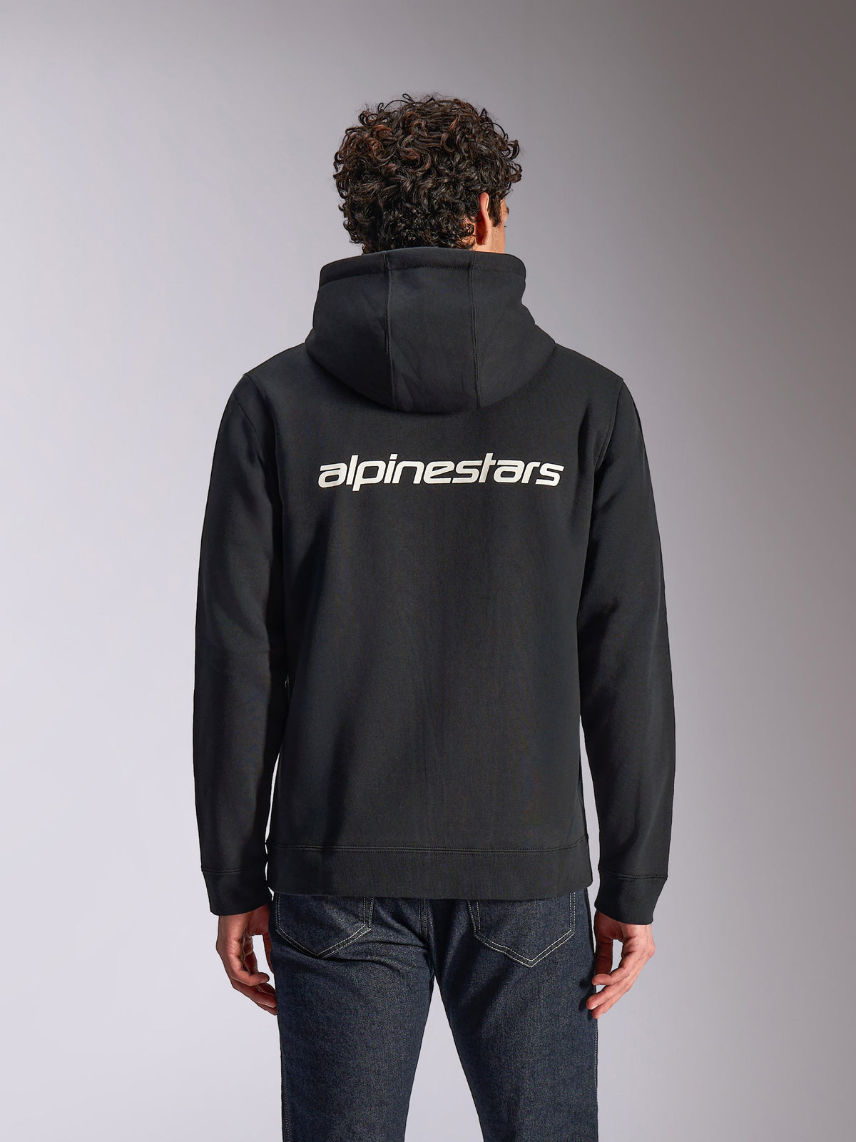 Alpinestars Transverse hættetrøje, afslappet streetwear, sort med hvidt Alpinestars-tekstlogo på ryggen, fleecehættetrøje med lynlås foran, lange ærmer, båret af en model med mørke denimjeans, moderne motorsport-inspireret design