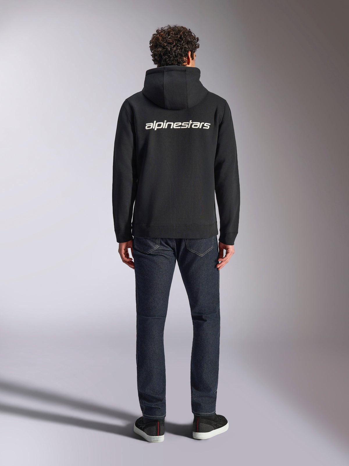 Alpinestars Transverse hættetrøje, casual motorsport streetwear, sort med hvidt Alpinestars-tekstlogo på ryggen, lange ærmer, ribkant forneden og ved manchetterne, set bagfra med mørke denimjeans