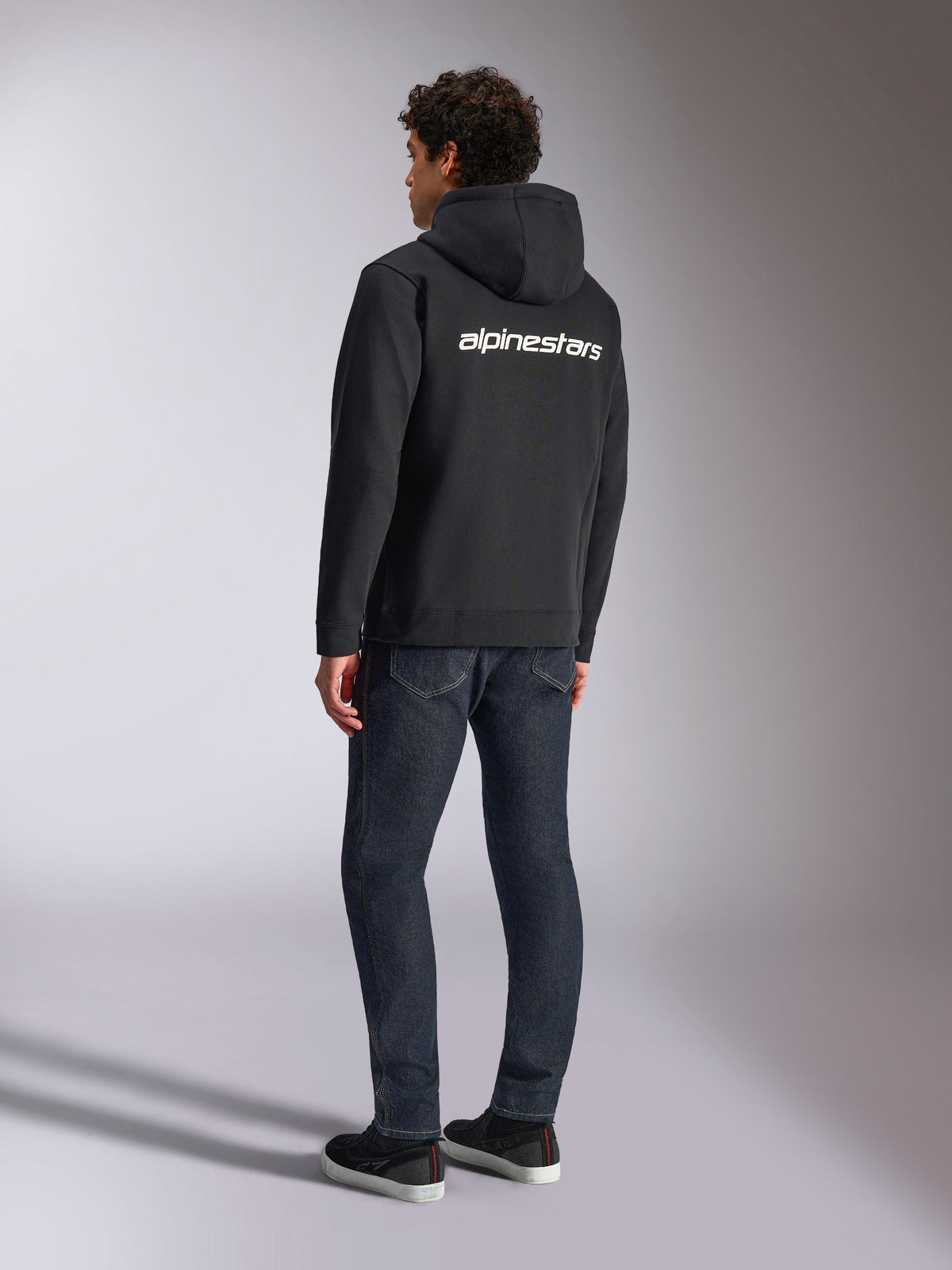 Alpinestars Transverse Hoodie, sort fleecehættetrøje med lynlås, set bagfra med hvidt Alpinestars-tekstlogo over skuldrene, båret med mørkeblå jeans og sorte sneakers, afslappet motorsport-inspireret streetwear