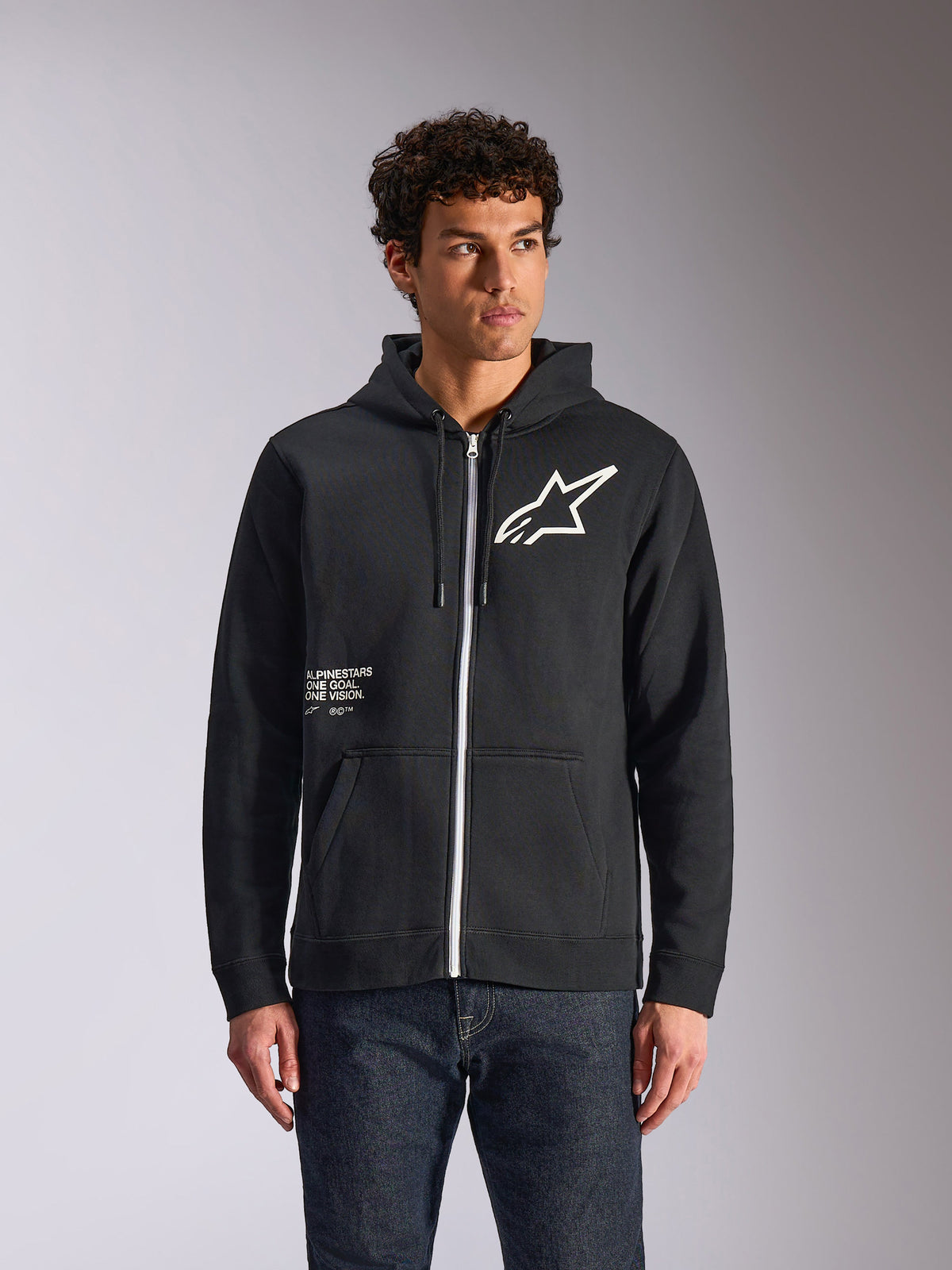 Alpinestars Transverse Hoodie, sort fleece-hættetrøje med lynlås foran, med hvidt Astars-logo og One Goal-grafik på forsiden, båret af en model med mørke jeans, afslappet motorsport-inspireret streetwear