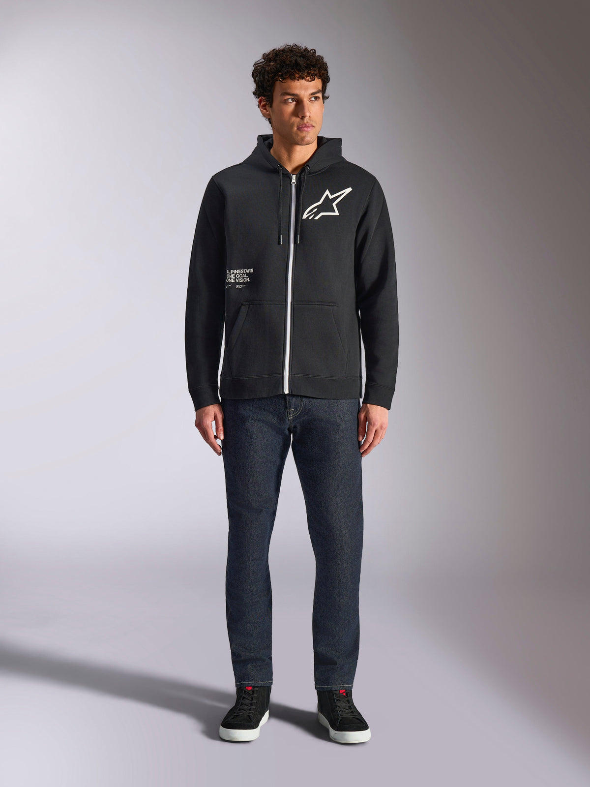 Alpinestars Transverse Hoodie, afslappet fleece med lynlås foran, sort, hvidt Astars-logo på brystet og 'One Goal'-print på forsiden, hætte med snøre, kombineret med mørke jeans og sorte sneakers, lifestyle motorsport streetwear