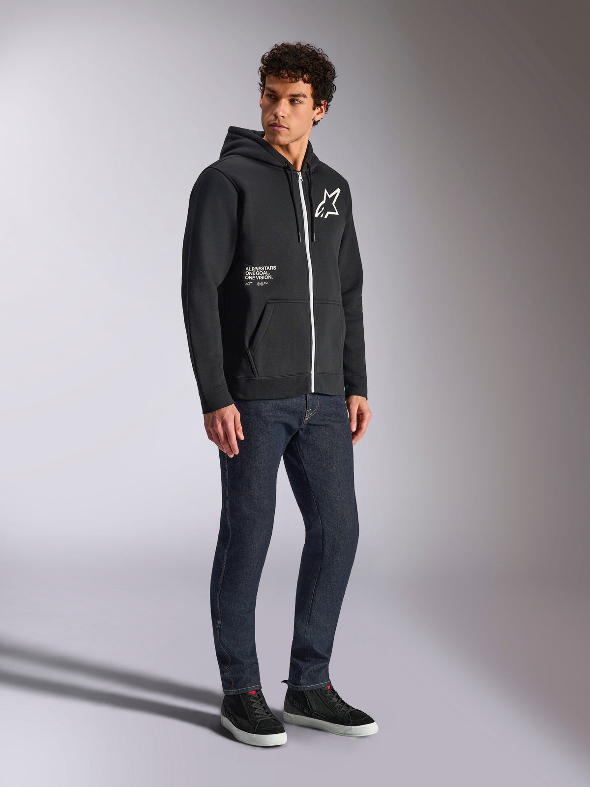 Alpinestars Transverse hættetrøje, sort hættetrøje med lynlås og hvid kontrastlynlås, med et hvidt stjernelogo på brystet og 'One Goal'-branding på lommen, båret af en model med mørke jeans og sorte sneakers, afslappet motorsport-inspireret streetwear.