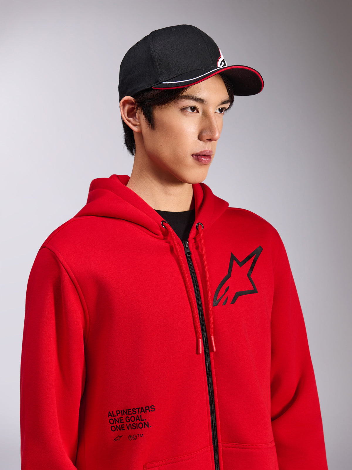 Alpinestars Transverse Hoodie, Red zip-front fleece, black Astars logo on chest and 'One Goal. One Vision.' text on the lower front, casual motorsport streetwear, worn by a model with a black cap