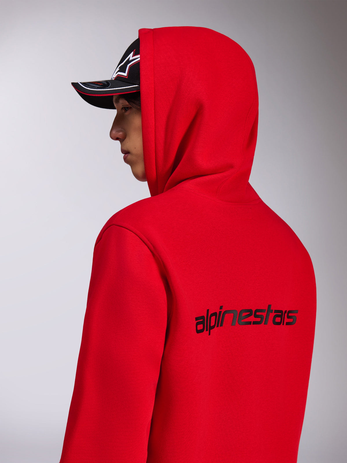 Alpinestars Transverse Hoodie, Red hooded fleece, side-back view showing the black Alpinestars wordmark on the back, worn with the hood up and a black cap, casual motorsport-inspired streetwear